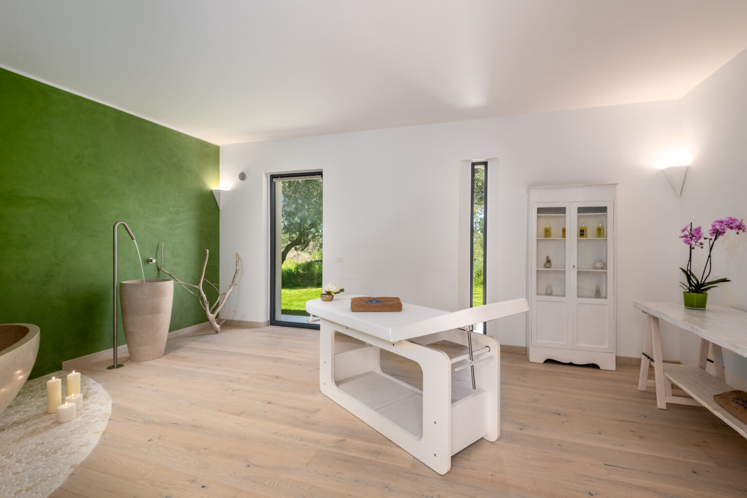 Modern spa interior featuring a calming green accent wall, a sleek treatment table, and natural light streaming through large windows. The space includes a minimalist decor with a bathtub, a glass cabinet, and potted orchids, creating a serene atmosphere for relaxation and wellness. | Only on SopranoVillas