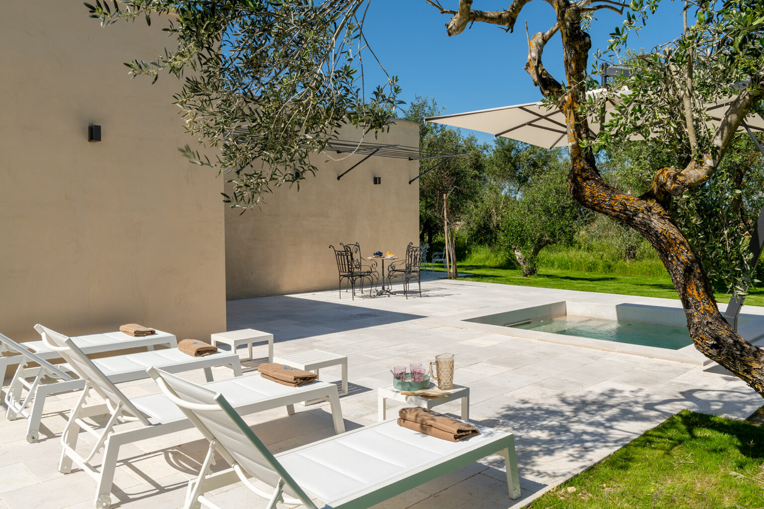 Modern outdoor patio featuring white lounge chairs, a small dining area, and a sleek pool surrounded by lush greenery and olive trees under a clear blue sky. Ideal for relaxation and leisure. | Only on SopranoVillas