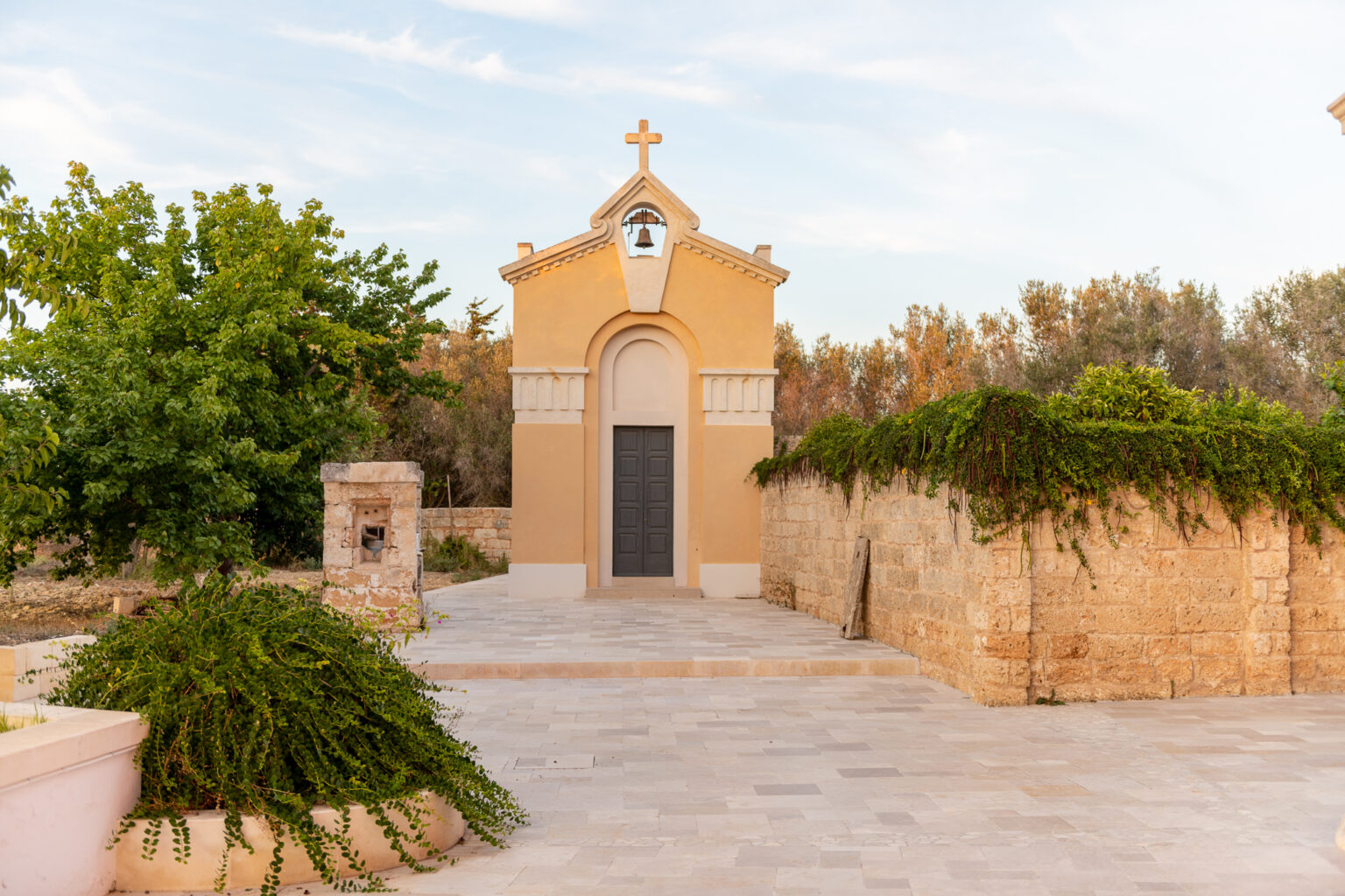 Small chapel surrounded by lush greenery and stone walls, set against a clear sky, showcasing traditional architecture and serene landscape. | Only on SopranoVillas
