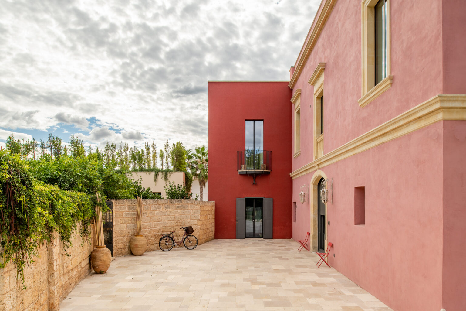 Modern architectural design featuring a vibrant red building with large windows, surrounded by greenery and a stone courtyard. A bicycle leans against the wall, adding a touch of charm to the outdoor space under a cloudy sky. | Only on SopranoVillas