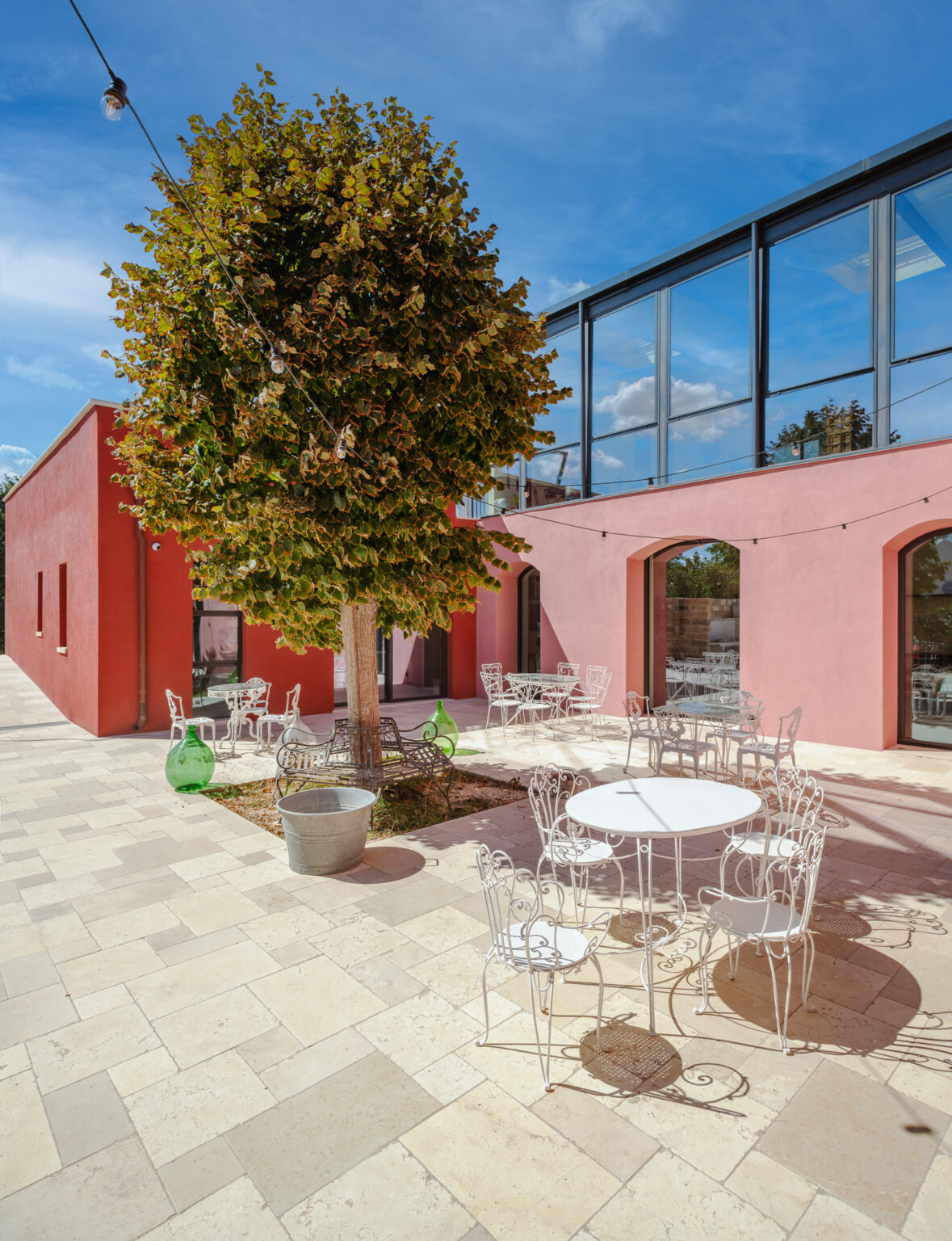Modern outdoor seating area with transparent chairs and a round table, surrounded by a vibrant red building and greenery under a clear blue sky. Ideal for casual dining or relaxation. | Only on SopranoVillas