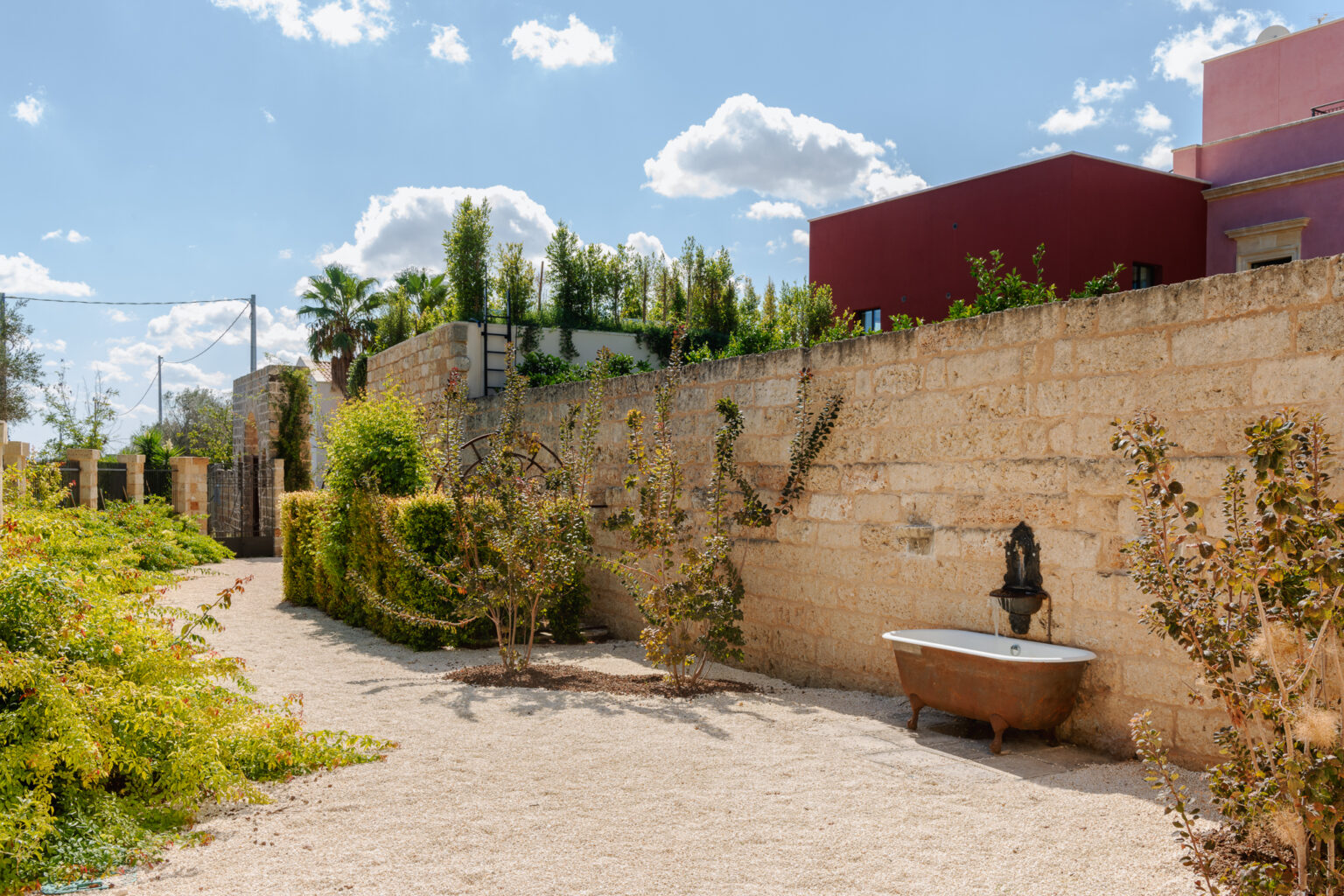 Charming outdoor pathway featuring a vintage bathtub and a stone wall adorned with greenery, under a bright blue sky with fluffy clouds. | Only on SopranoVillas