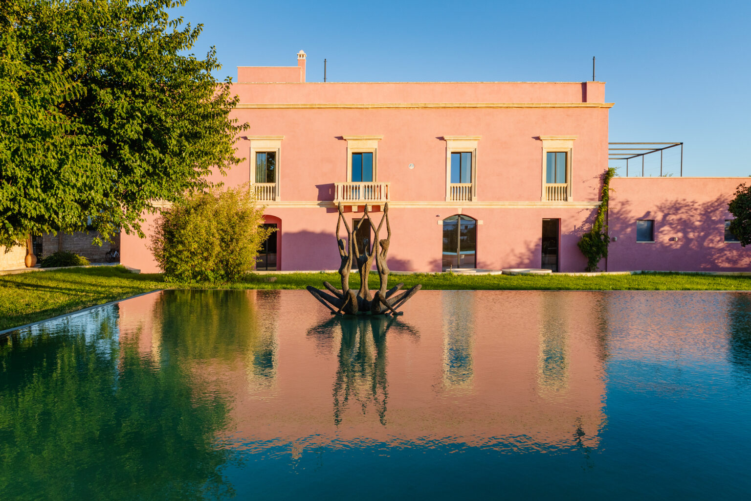A serene outdoor scene featuring a pink building reflecting in a tranquil pool, complemented by a striking sculpture of figures holding hands at the center. Lush greenery surrounds the area, enhancing the peaceful atmosphere. | Only on SopranoVillas