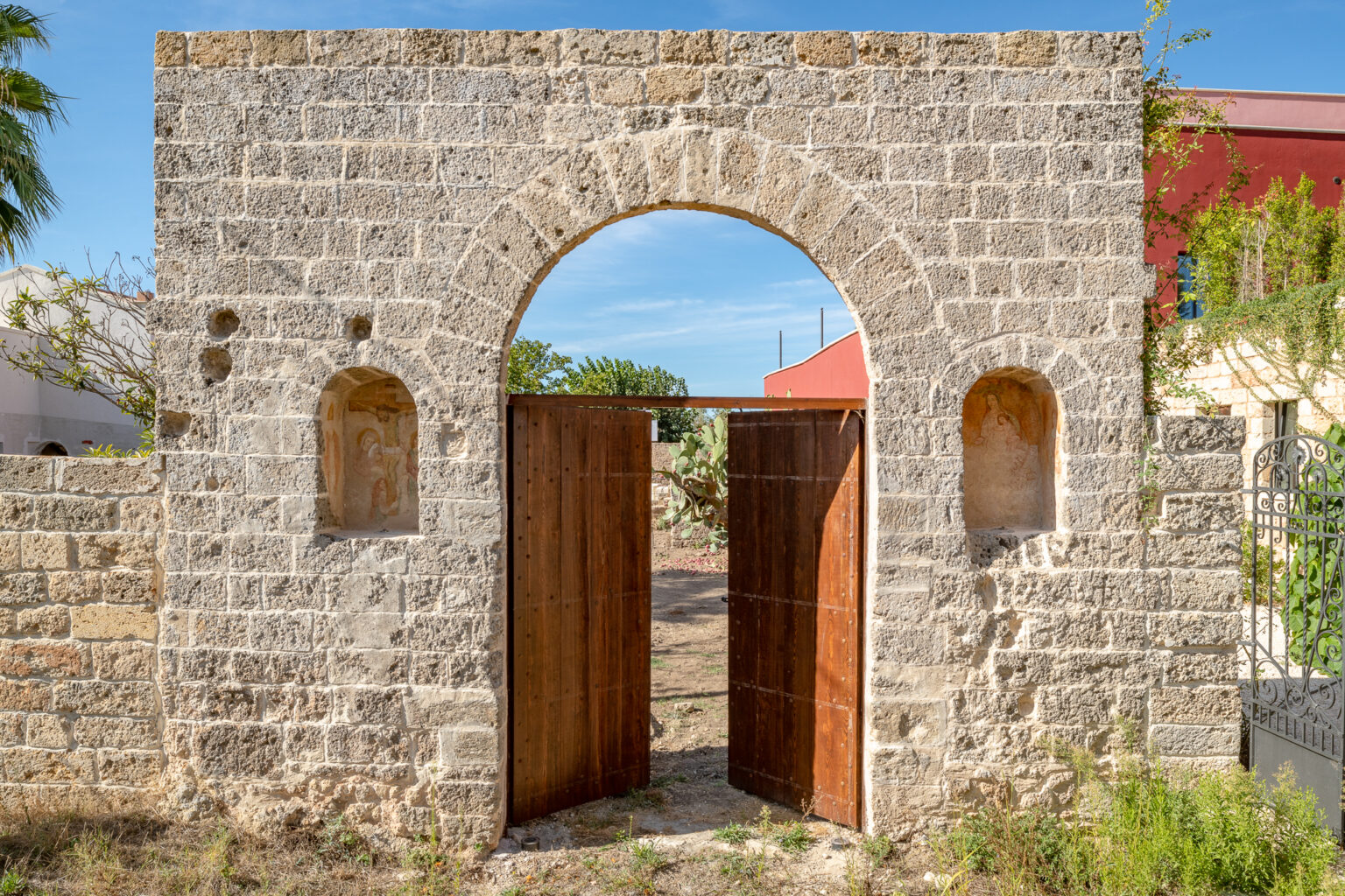 Stone archway entrance with wooden doors, framed by a textured stone wall featuring decorative niches, surrounded by greenery and a blue sky. Ideal for showcasing historical architecture and rustic design. | Only on SopranoVillas
