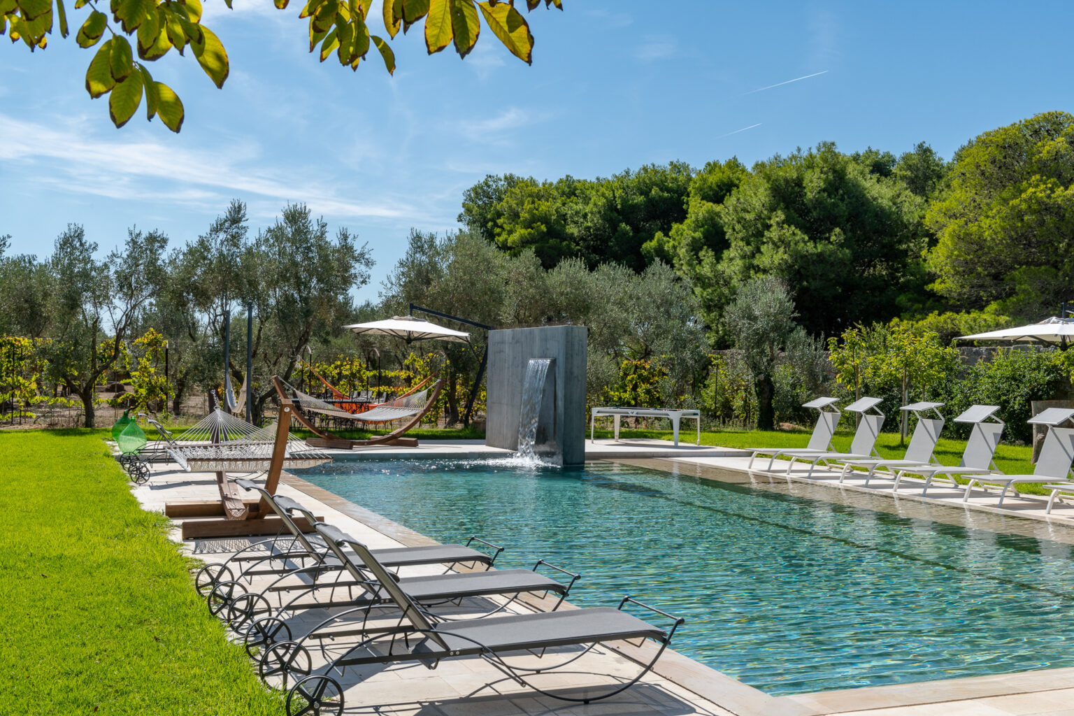 Luxurious outdoor pool area featuring sun loungers, hammocks, and a modern water feature, surrounded by lush greenery and olive trees under a clear blue sky. Perfect for relaxation and leisure in a serene garden setting. | Only on SopranoVillas