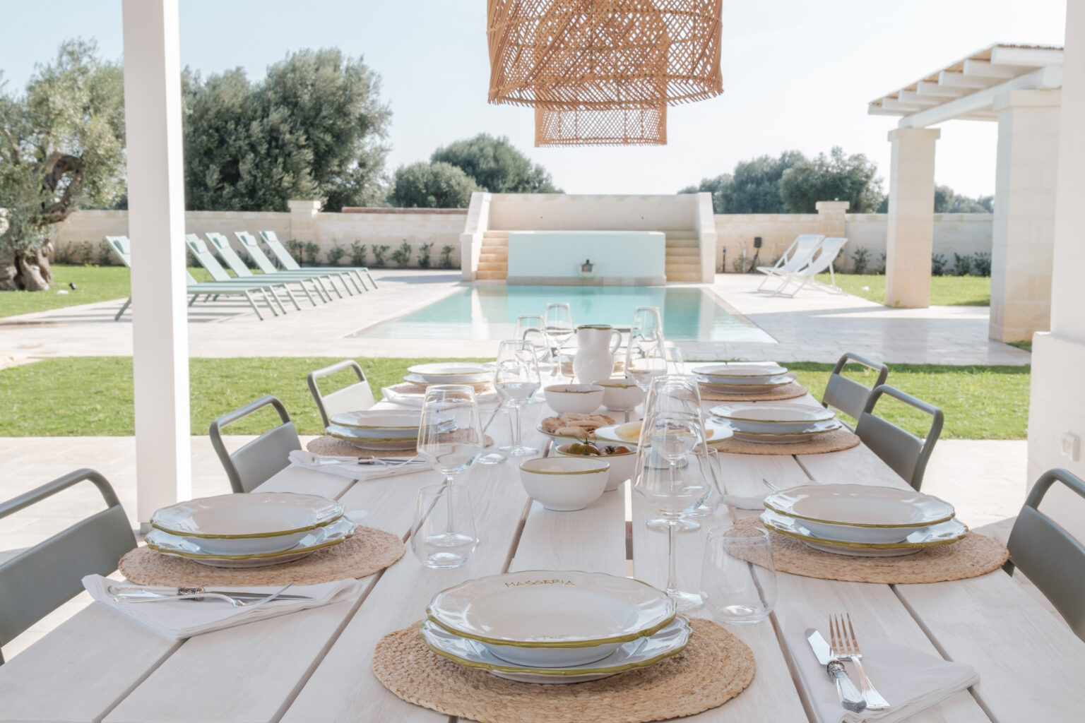 Elegant outdoor dining setup featuring a long wooden table adorned with decorative plates, glassware, and woven placemats, set against a backdrop of a serene swimming pool and olive trees. Ideal for alfresco dining experiences in a luxurious villa setting. | Only on SopranoVillas