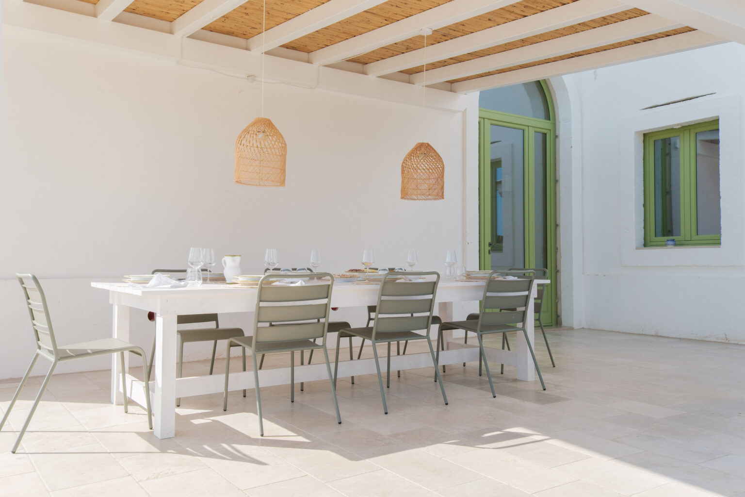 Bright and airy outdoor dining area featuring a white wooden table set with glassware and plates, surrounded by modern green chairs. Two woven pendant lights hang from a wooden ceiling, enhancing the inviting atmosphere. The space is accentuated by light-colored stone flooring and vibrant green window frames, ideal for alfresco dining. | Only on SopranoVillas