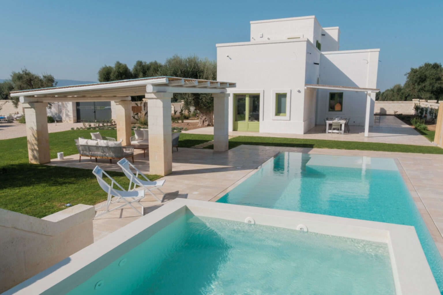 Modern villa with two swimming pools, outdoor seating area, and lush green lawn under a clear blue sky. Ideal for luxury vacations and relaxation. | Only on SopranoVillas
