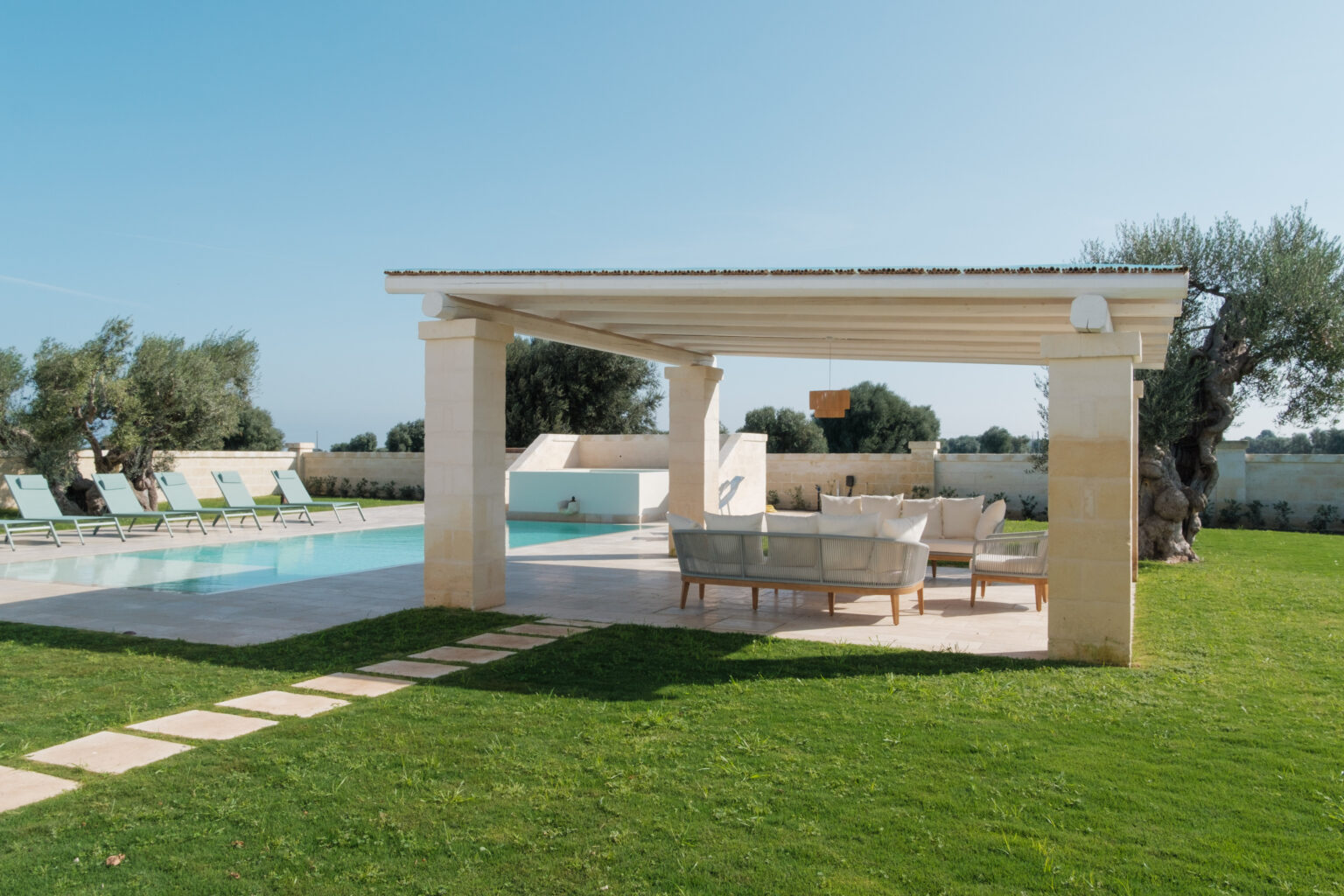 Modern outdoor living space featuring a swimming pool, stylish lounge area with comfortable seating, and lush green lawn, surrounded by olive trees under a clear blue sky. | Only on SopranoVillas