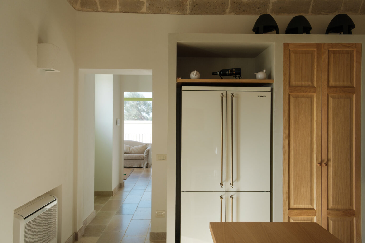 Modern kitchen interior featuring a sleek white SMEG refrigerator, wooden cabinets, and a view into a cozy living area. Natural light floods the space, highlighting the minimalist design and stone flooring. | Only on SopranoVillas