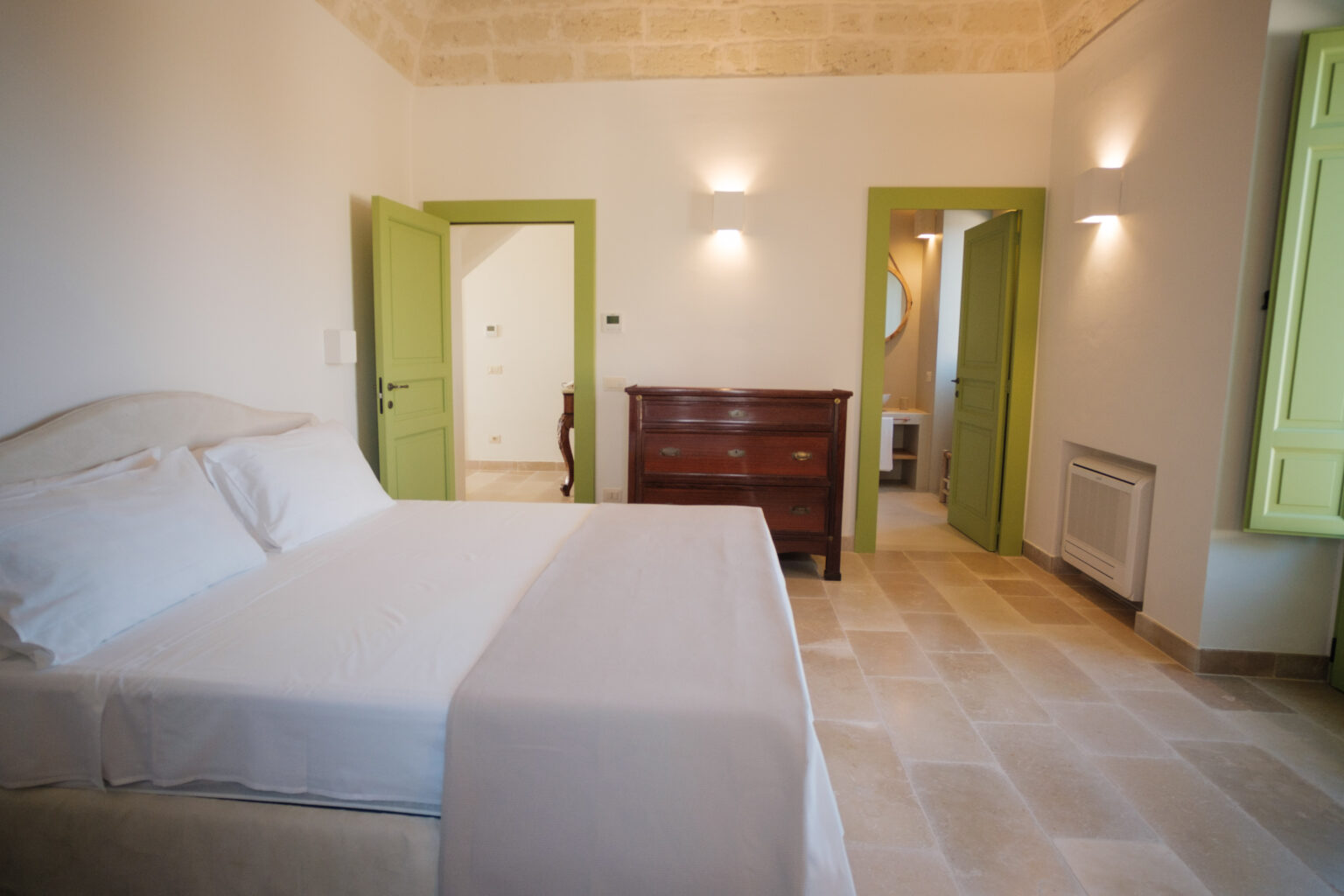 Cozy bedroom featuring a double bed with white linens, light-colored stone flooring, and green accent doors. The room is well-lit with modern wall sconces, showcasing a warm and inviting atmosphere, perfect for relaxation. | Only on SopranoVillas