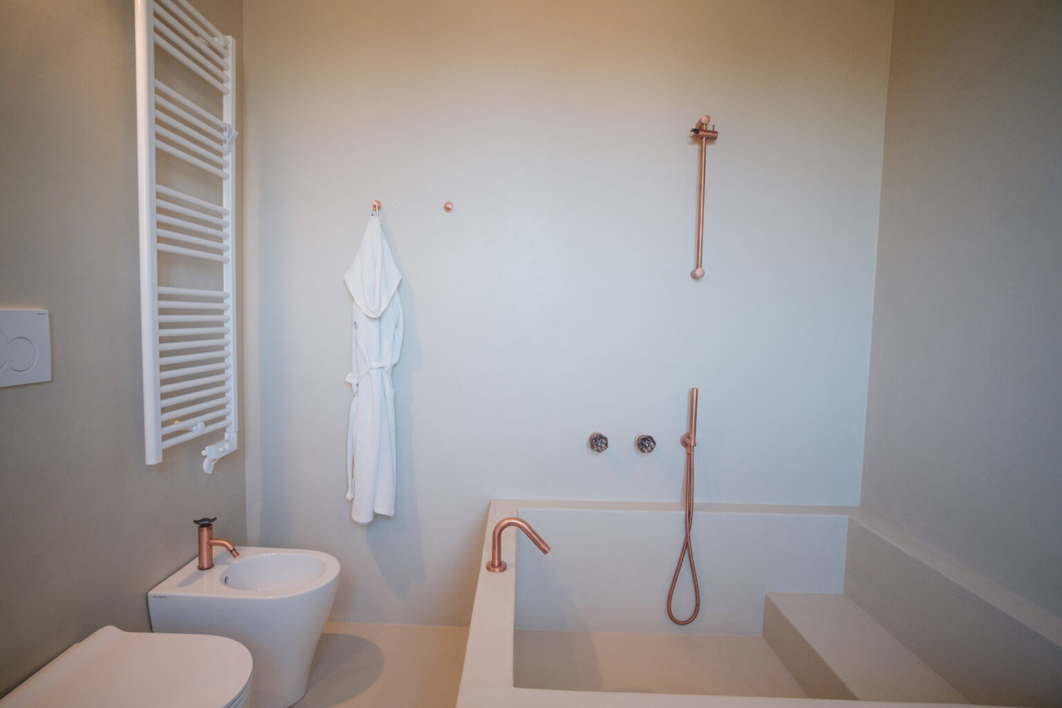 Modern bathroom featuring a minimalist design with a freestanding bathtub, a wall-mounted shower fixture, and a bidet. The space is accented with rose gold fixtures and a white bathrobe hanging on the wall, showcasing a clean and contemporary aesthetic. | Only on SopranoVillas