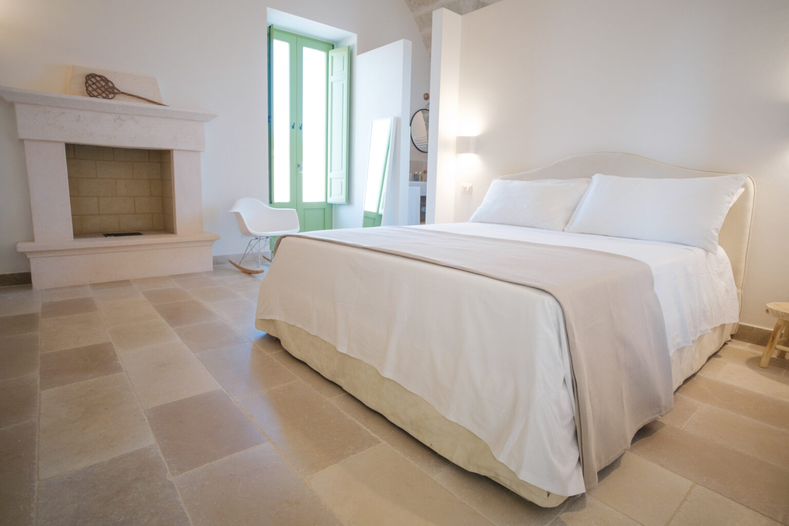 Cozy bedroom featuring a white bed with plush pillows, a minimalist rocking chair, and a stone fireplace. Large green windows allow natural light to brighten the space, complemented by light-colored stone flooring. Ideal for relaxation and comfort. | Only on SopranoVillas