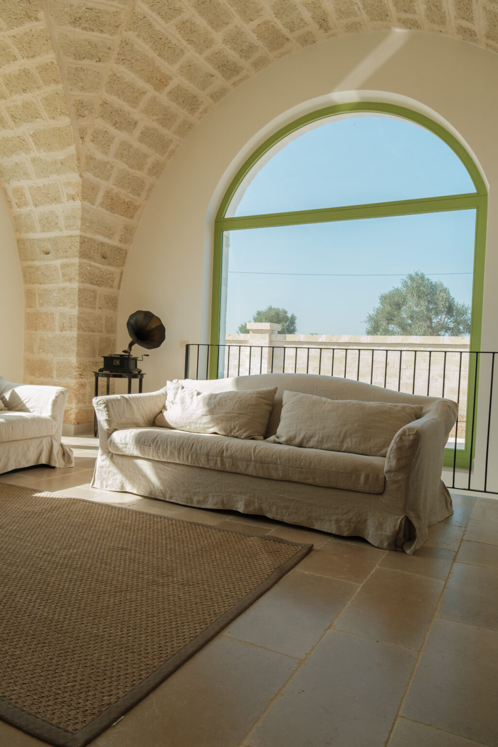 Contemporary living room featuring a light-colored sofa with cushions, a woven rug on stone flooring, and a large arched window with green frames allowing natural light. A vintage gramophone adds a touch of elegance to the space. | Only on SopranoVillas