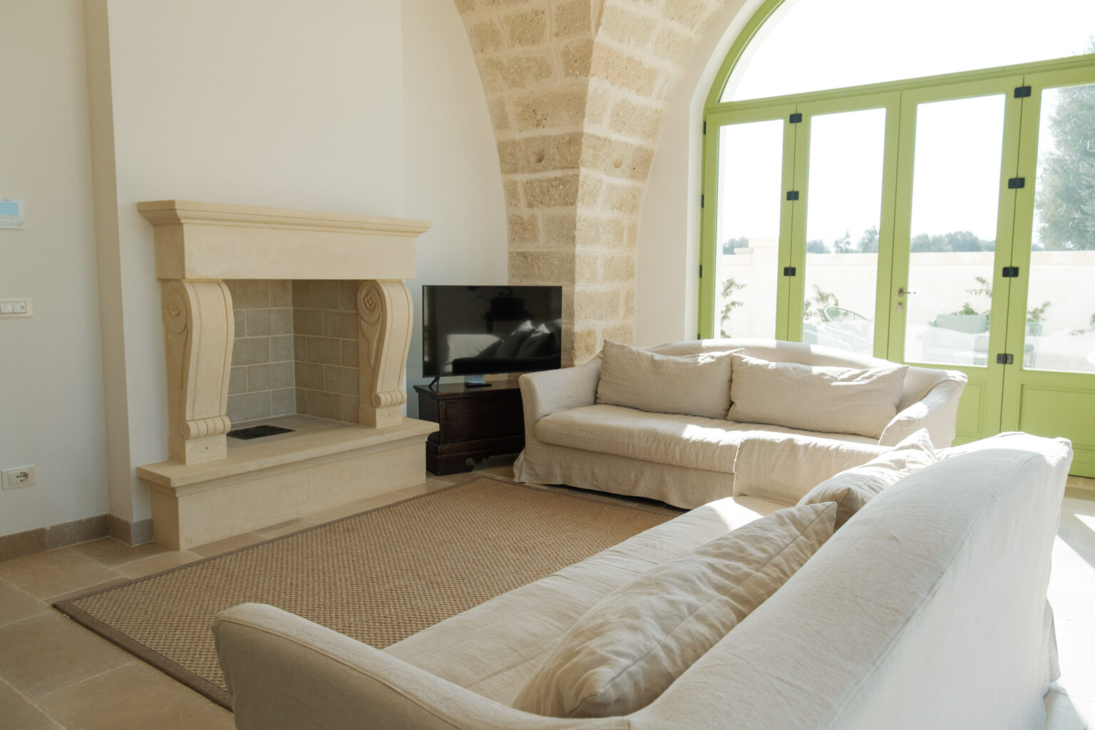Bright and airy living room featuring a stone fireplace, beige sofas, and large green windows. The space is adorned with natural light, creating a warm and inviting atmosphere. Ideal for relaxation and social gatherings. | Only on SopranoVillas