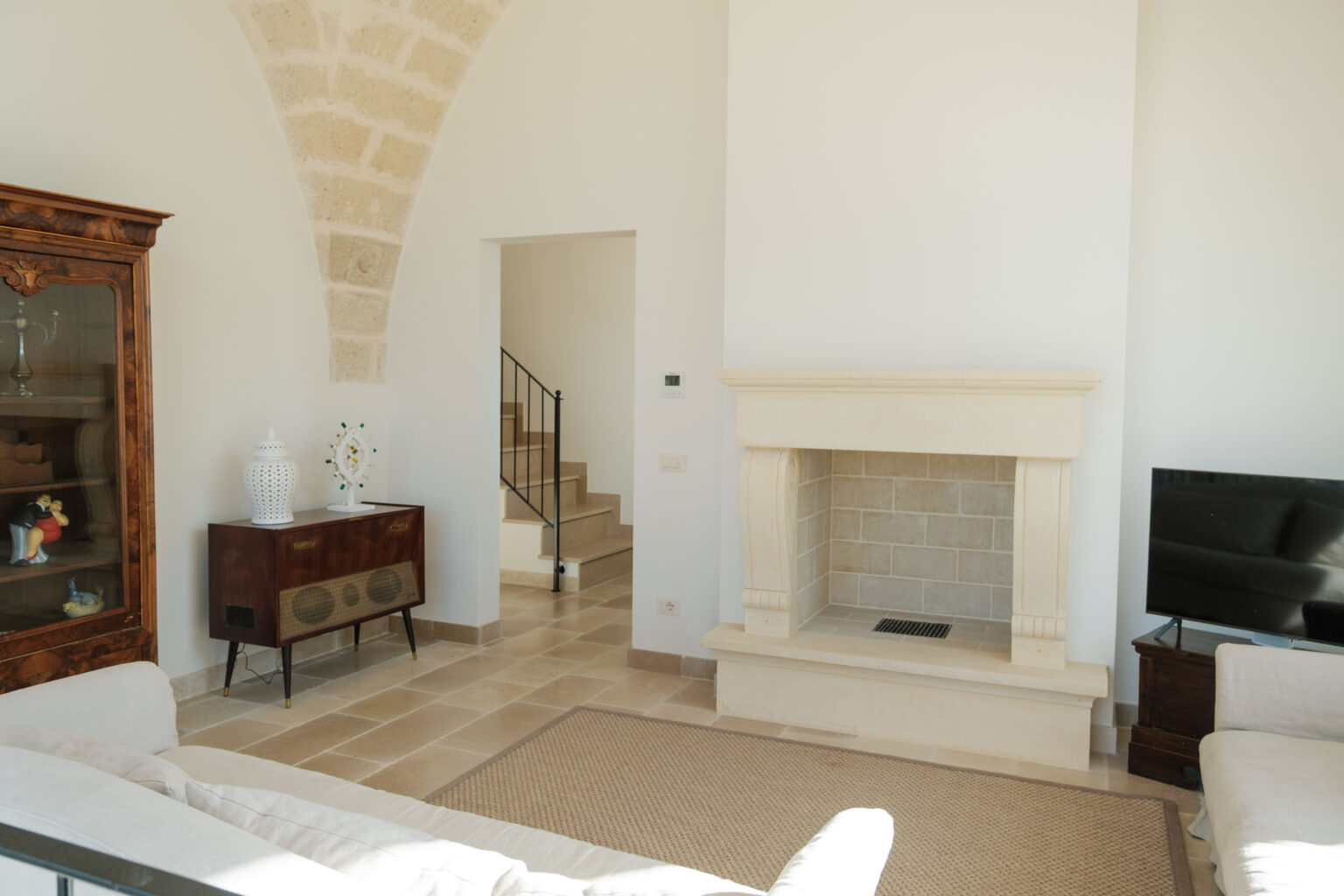 Bright and airy living room featuring a stone fireplace, modern furniture, and a staircase leading to the upper level. The room showcases a blend of contemporary and classic decor with a wooden cabinet and decorative accents. Natural light illuminates the space, highlighting the neutral color palette and elegant design elements. | Only on SopranoVillas