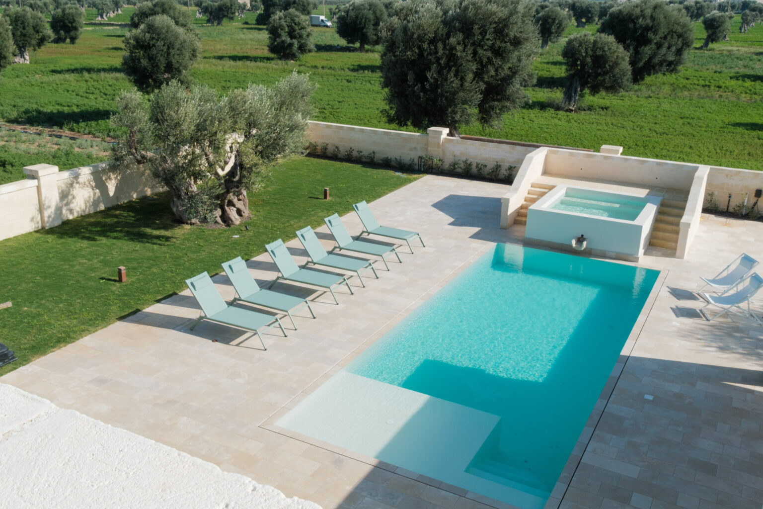 Aerial view of a luxurious outdoor pool area featuring a sleek swimming pool, a hot tub, and several modern lounge chairs on a stone patio, surrounded by lush green fields and olive trees, ideal for relaxation and leisure. | Only on SopranoVillas