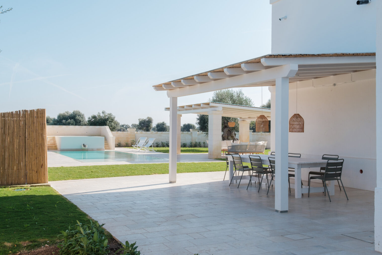 Modern outdoor living space featuring a white patio with a dining table, overlooking a serene swimming pool surrounded by lush greenery and olive trees. Ideal for relaxation and entertaining in a sunny environment. | Only on SopranoVillas