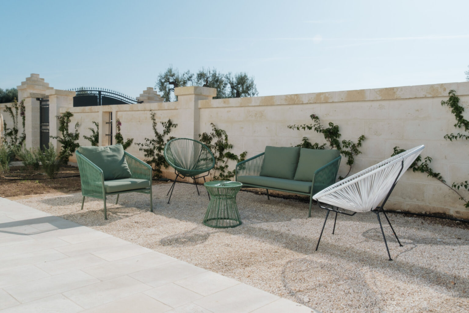 Outdoor seating area featuring modern green and white lounge chairs and a small table on a gravel surface, surrounded by a stone wall and climbing plants under a clear blue sky. | Only on SopranoVillas
