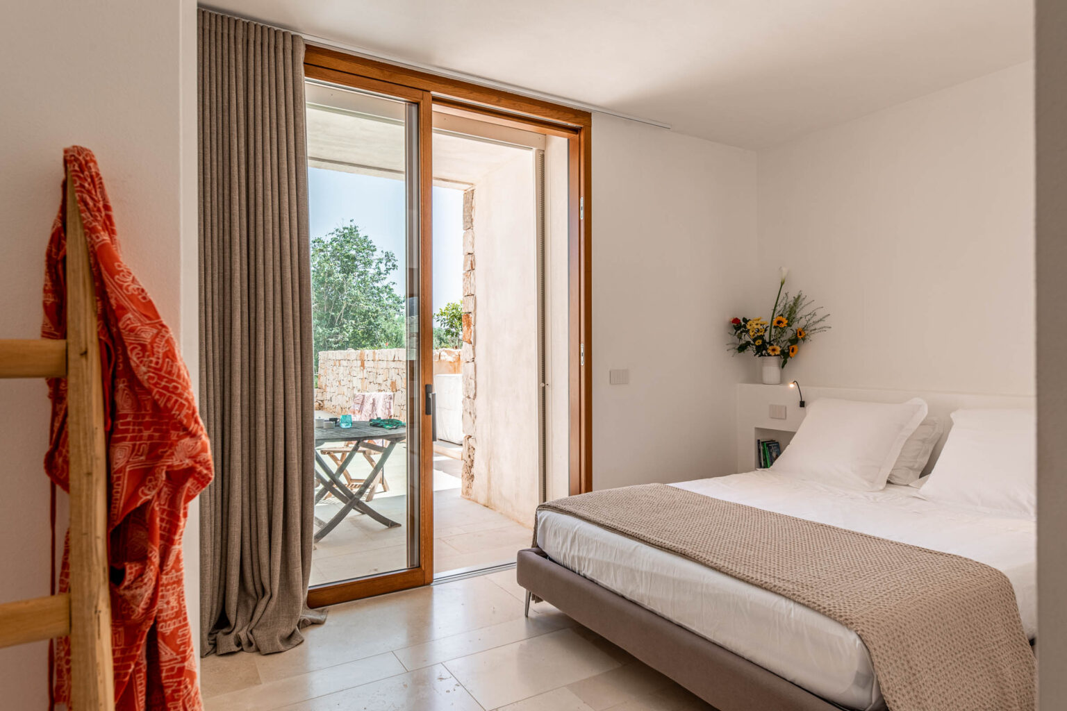 Modern bedroom with a double bed, neutral-toned bedding, and a cozy atmosphere. Features large glass doors leading to a balcony with outdoor seating, natural light, and stylish decor including a floral arrangement and a warm throw. Ideal for relaxation and comfort. | Only on SopranoVillas