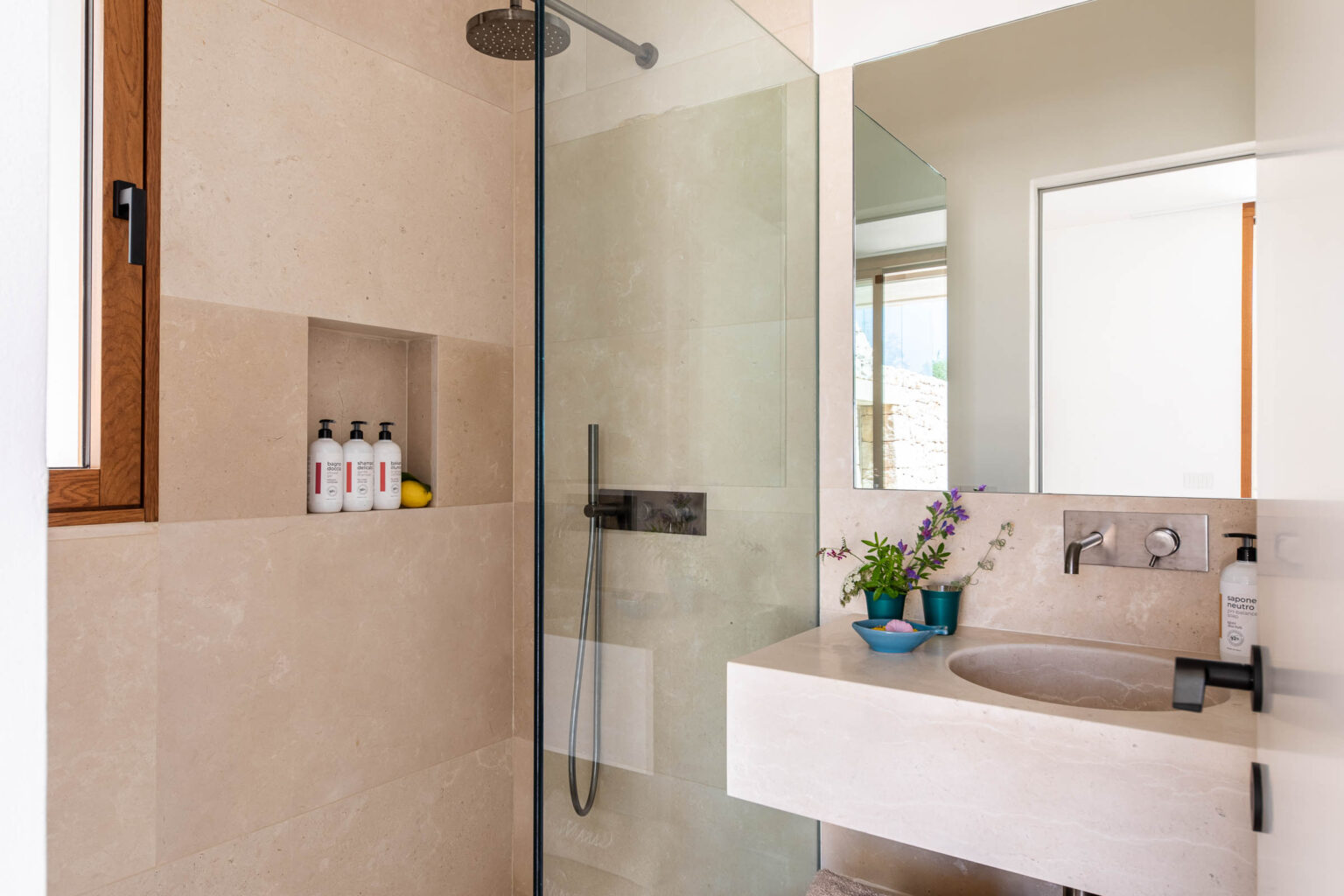 Modern bathroom featuring a walk-in shower with glass doors, a sleek sink with a minimalist design, and natural light streaming through a window. Decorative elements include small bottles on a shelf and a potted plant, highlighting a clean and stylish aesthetic. | Only on SopranoVillas