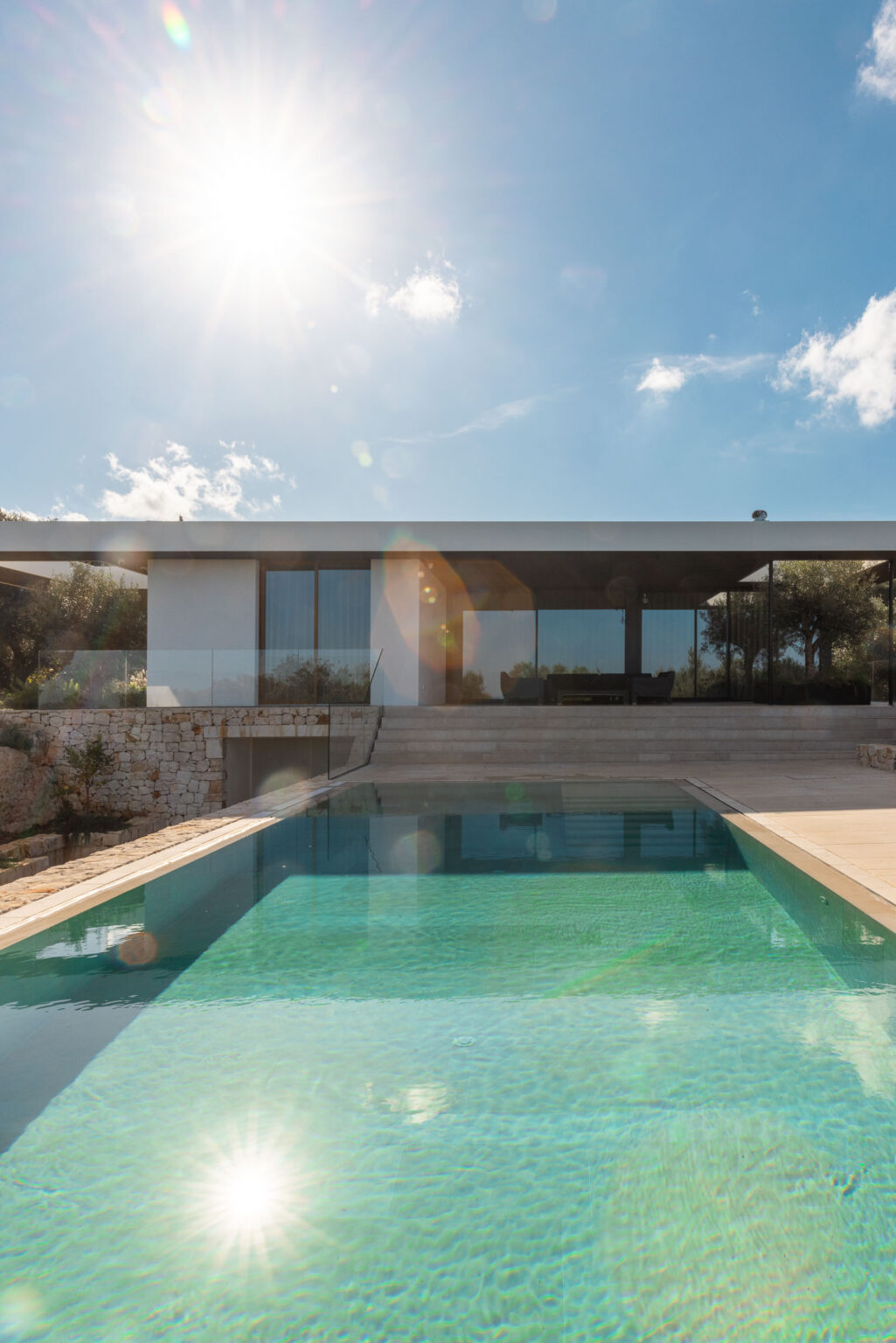 Modern villa with a sleek design featuring large glass windows, surrounded by greenery and a swimming pool in the foreground, under a bright blue sky. | Only on SopranoVillas