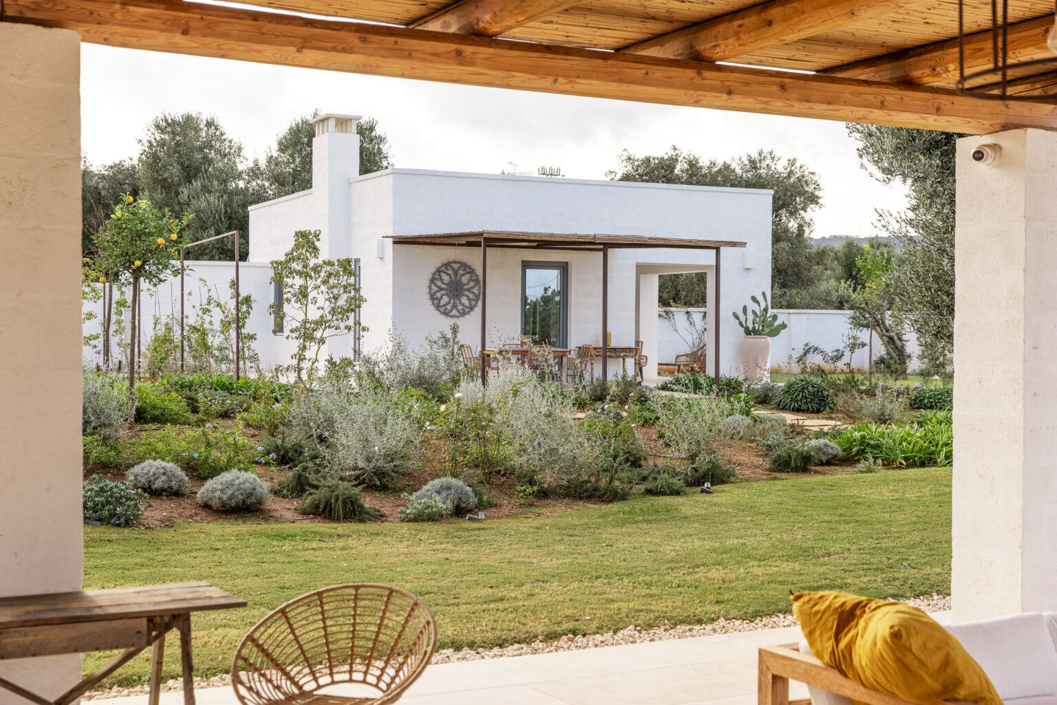 Modern white house surrounded by a lush garden, viewed from a patio with outdoor seating. The landscape features various plants and trees, creating a serene outdoor atmosphere. | Only on SopranoVillas
