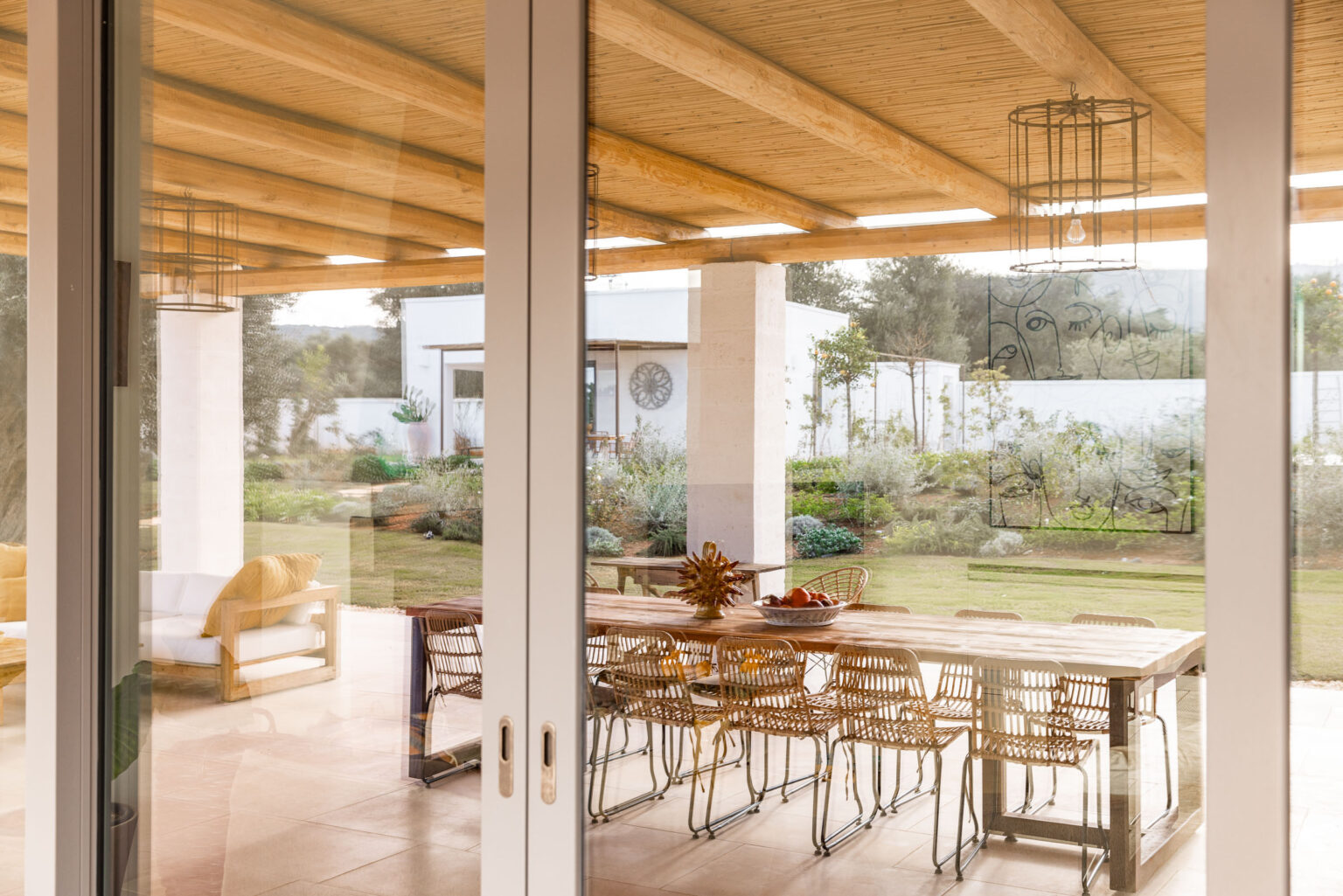 Modern outdoor dining area with a large wooden table and stylish chairs, surrounded by a lush garden and natural light pouring in through large glass doors. | Only on SopranoVillas