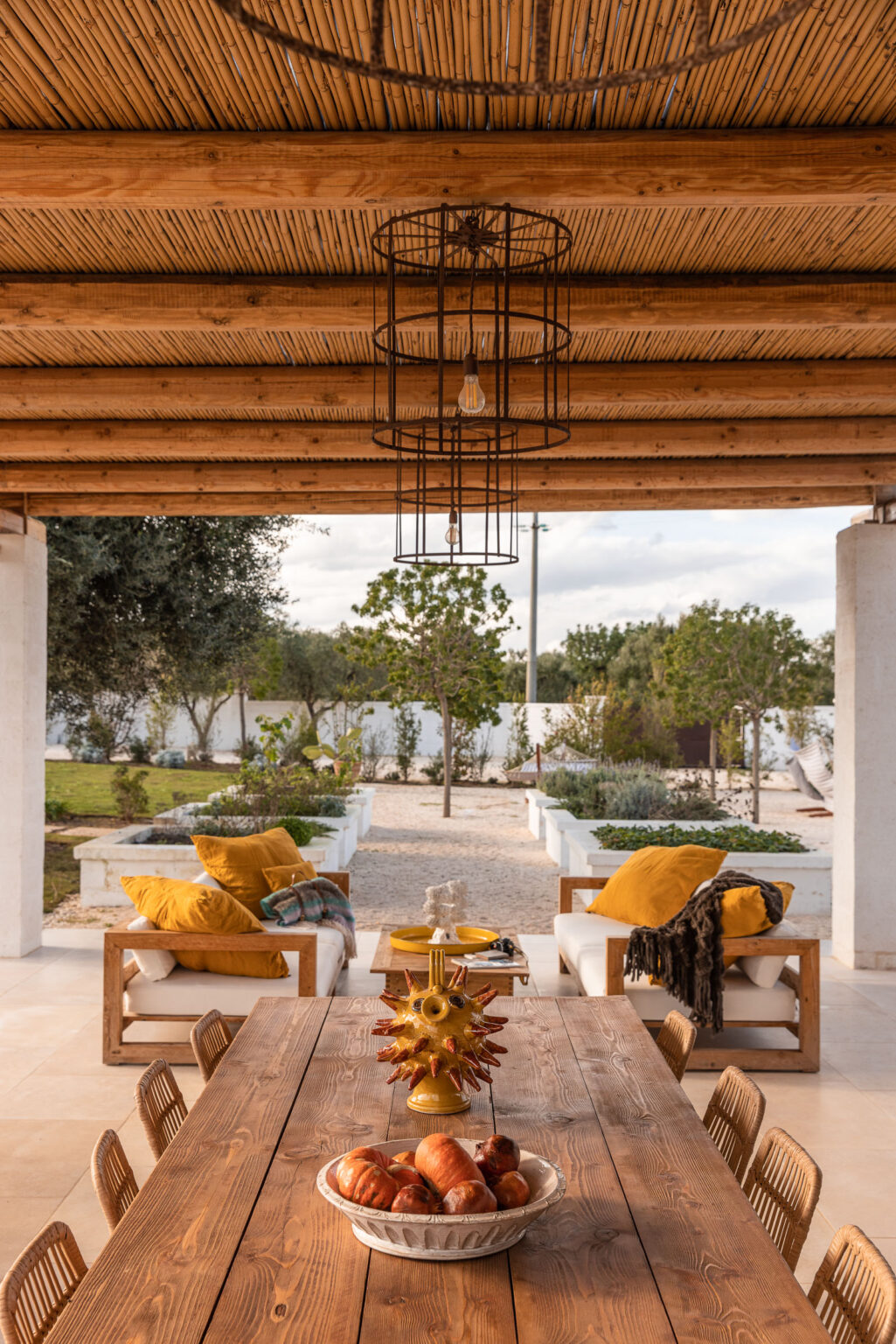 Cozy outdoor seating area with yellow cushions, surrounded by lush greenery and a gravel pathway, under a wooden pergola with a stylish hanging light fixture. Ideal for relaxation and entertaining in a tranquil garden setting. | Only on SopranoVillas