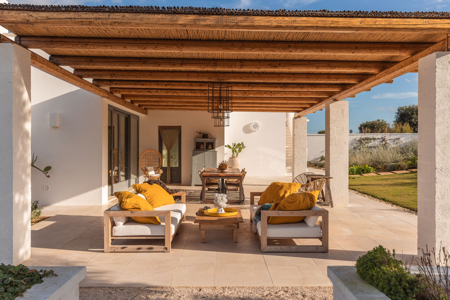 Spacious outdoor patio featuring wooden beams, comfortable seating with yellow cushions, and a dining table, surrounded by a lush garden and clear blue sky. | Only on SopranoVillas