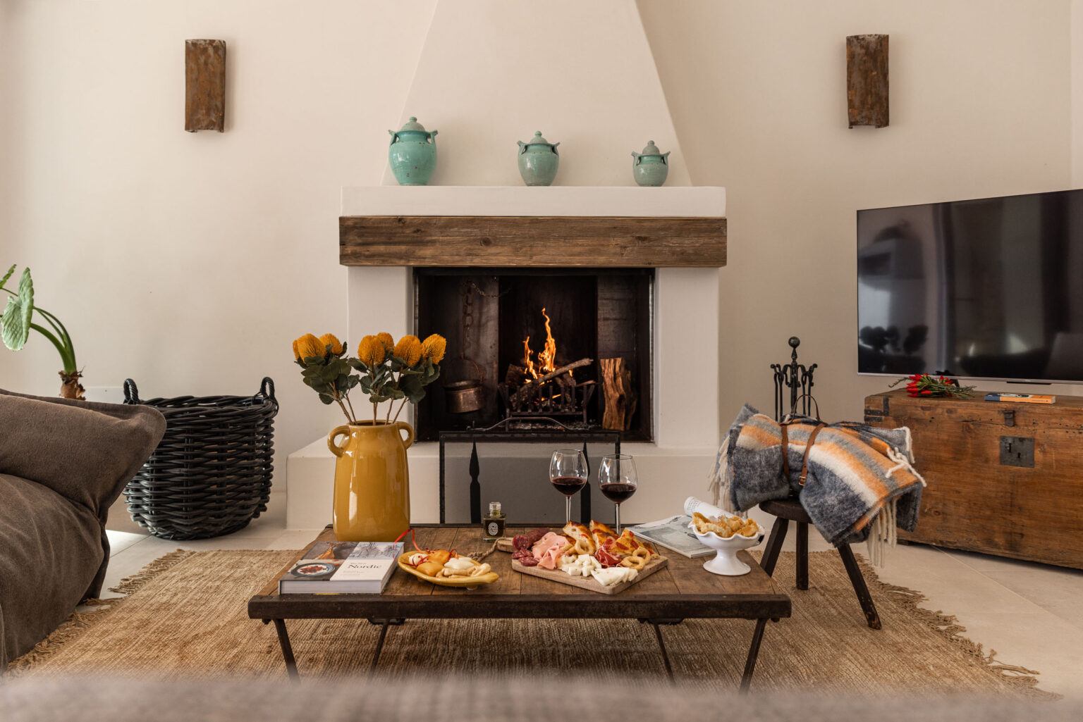 Cozy living room featuring a rustic fireplace, wooden coffee table with a charcuterie spread, wine glasses, and a vase of flowers, complemented by a comfortable sofa and modern decor. | Only on SopranoVillas