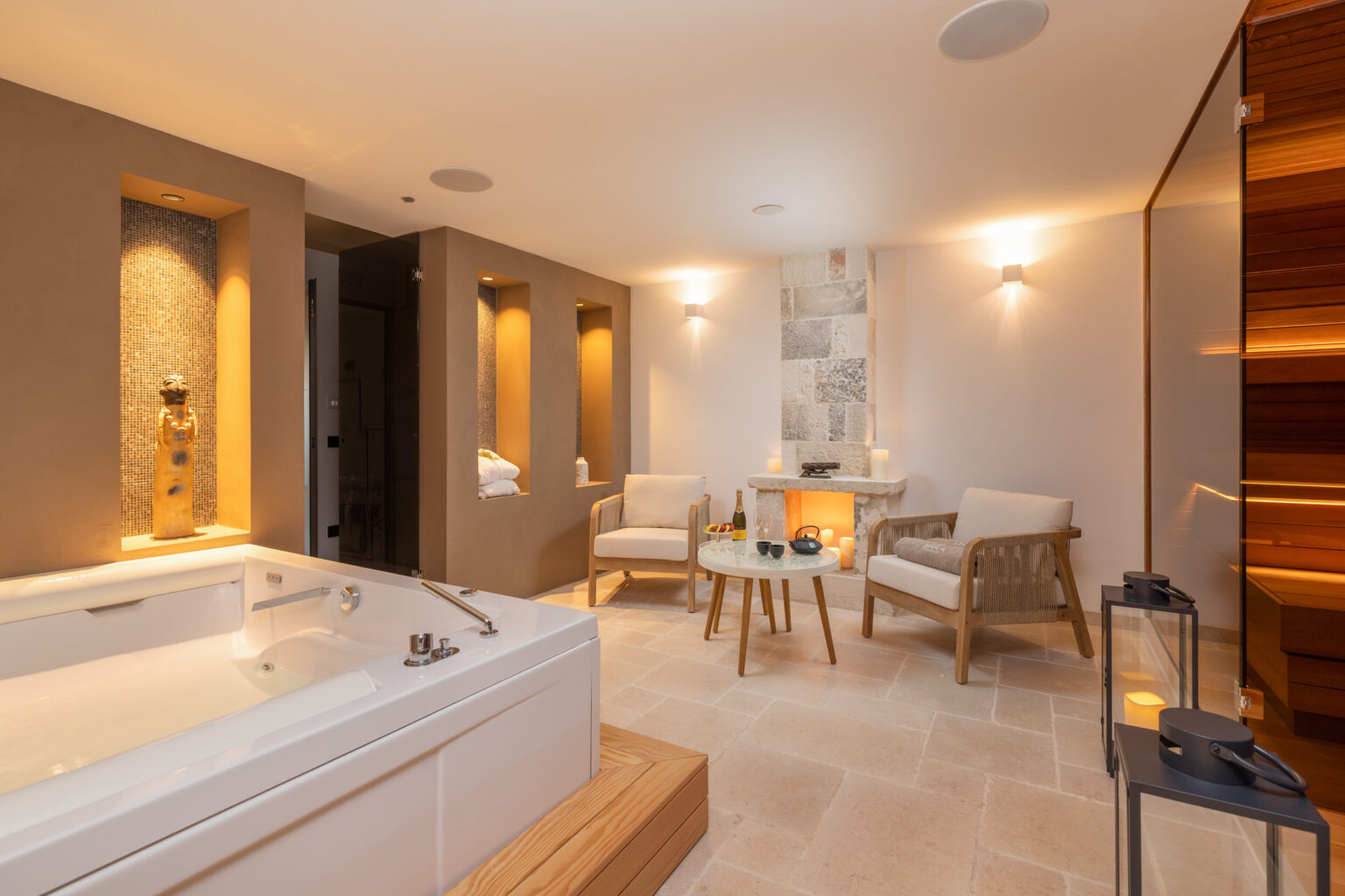 Luxurious spa interior featuring a white jacuzzi, elegant seating area with wooden chairs, a stone fireplace, and ambient lighting. The serene space is designed for relaxation and wellness, complemented by decorative elements and soft textiles. | Only on SopranoVillas