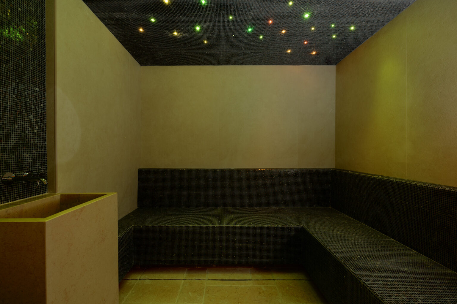 Interior view of a modern steam room featuring dark stone seating, a decorative wall fountain, and a ceiling adorned with colorful, twinkling lights, creating a relaxing atmosphere. | Only on SopranoVillas