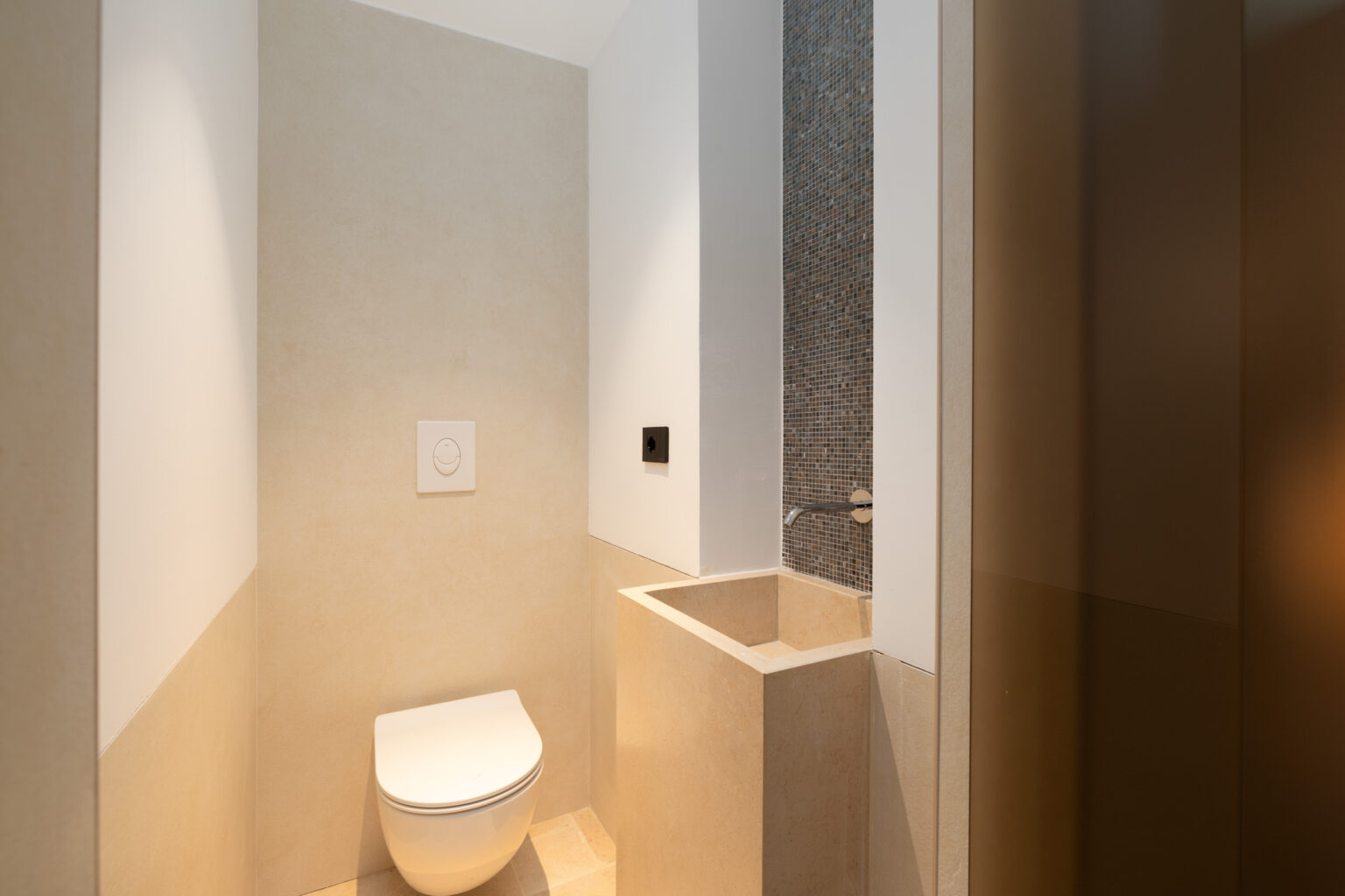 Modern bathroom interior featuring a minimalist design with a wall-mounted toilet and a sleek sink area. The walls are finished in light neutral tones, complemented by a textured stone backsplash. Ideal for showcasing contemporary bathroom aesthetics. | Only on SopranoVillas