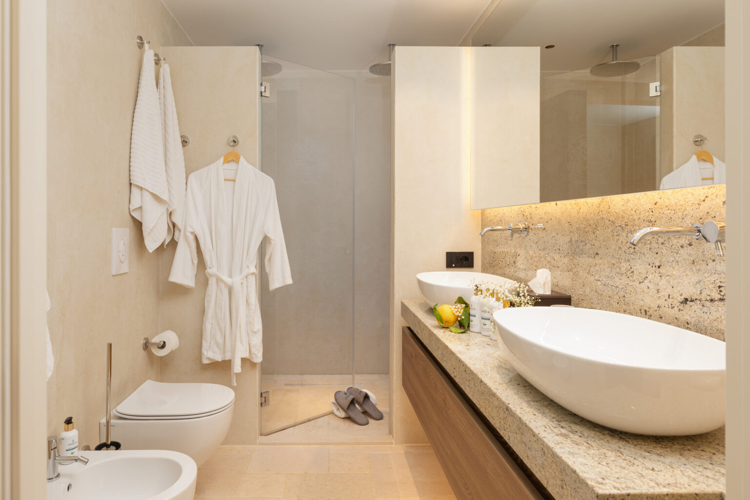 Modern bathroom featuring a spacious layout with a large oval sink, elegant countertops, a walk-in shower, and plush white robes hanging on the wall. The design emphasizes luxury and comfort, with neutral tones and soft lighting. | Only on SopranoVillas