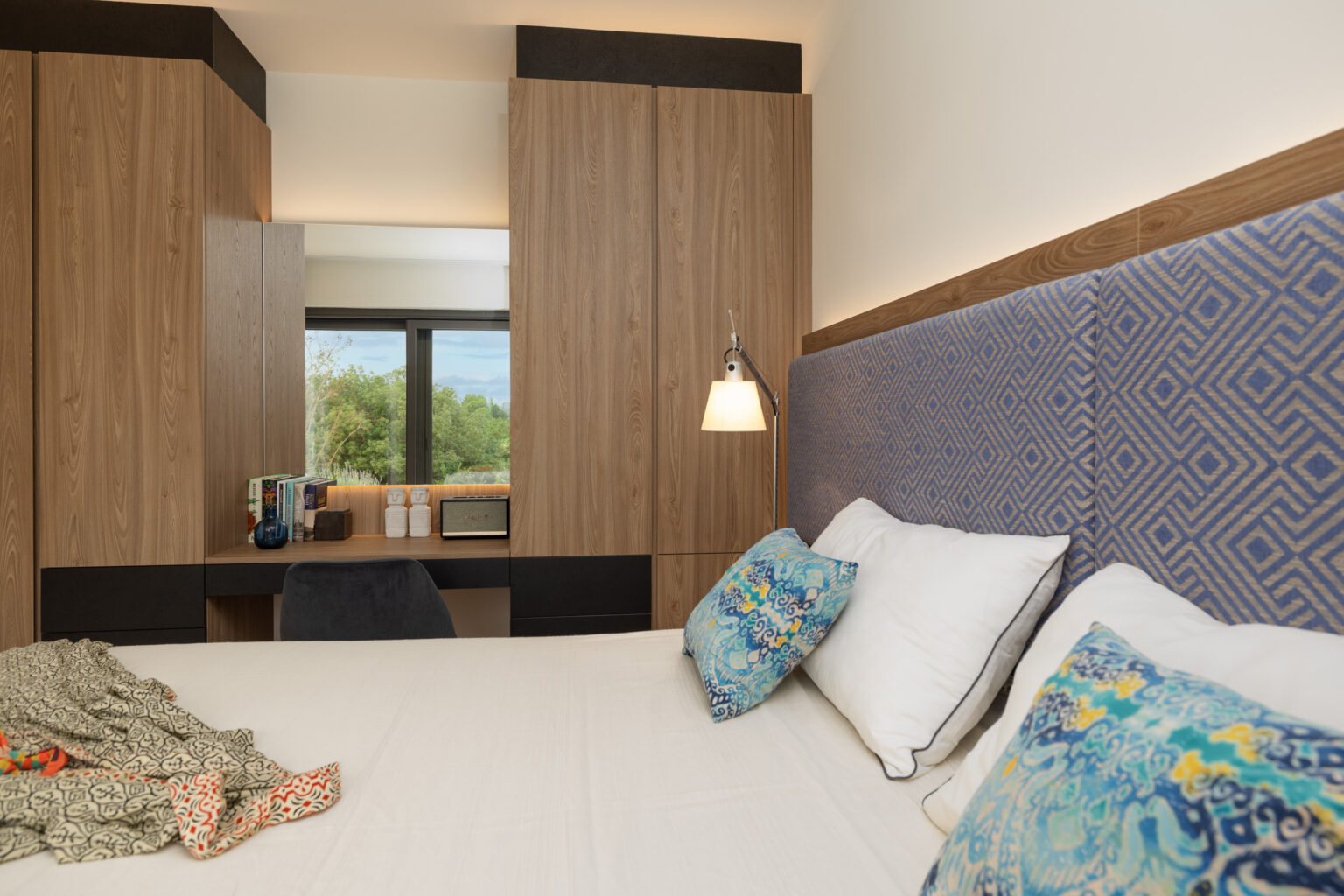 Modern hotel room featuring a comfortable bed with decorative pillows, stylish wooden cabinetry, and a cozy workspace. Natural light flows in from large windows, enhancing the inviting atmosphere. | Only on SopranoVillas