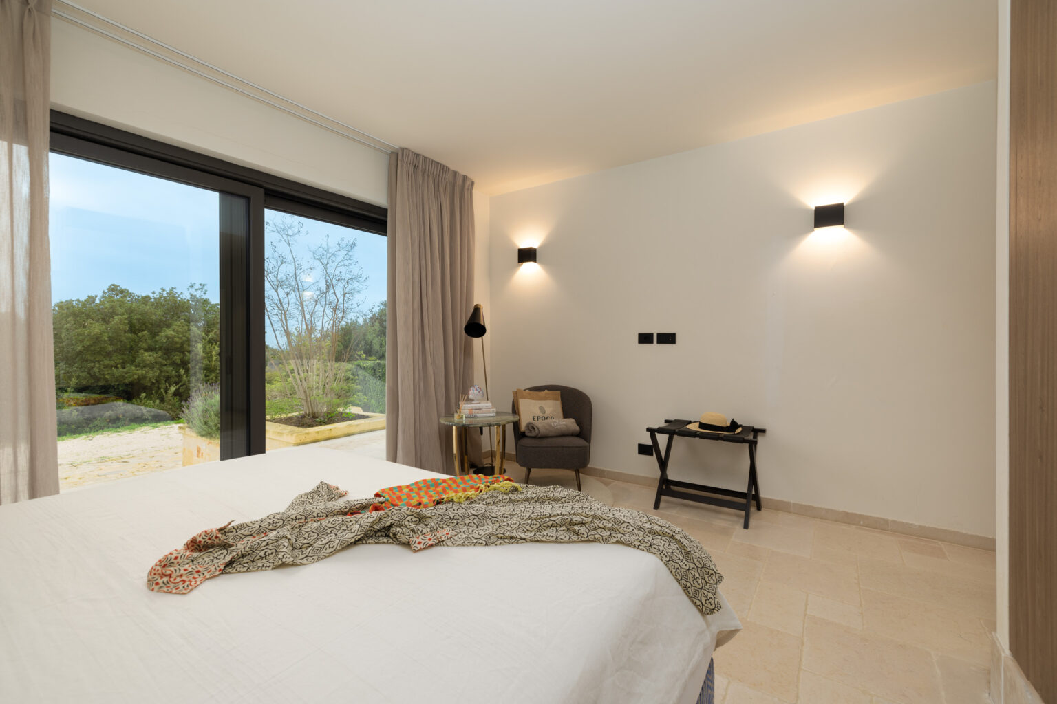 Modern bedroom interior featuring a cozy bed with a decorative throw, a stylish armchair, and a small side table. Large windows provide natural light and a view of the outdoors, enhancing the serene atmosphere. | Only on SopranoVillas