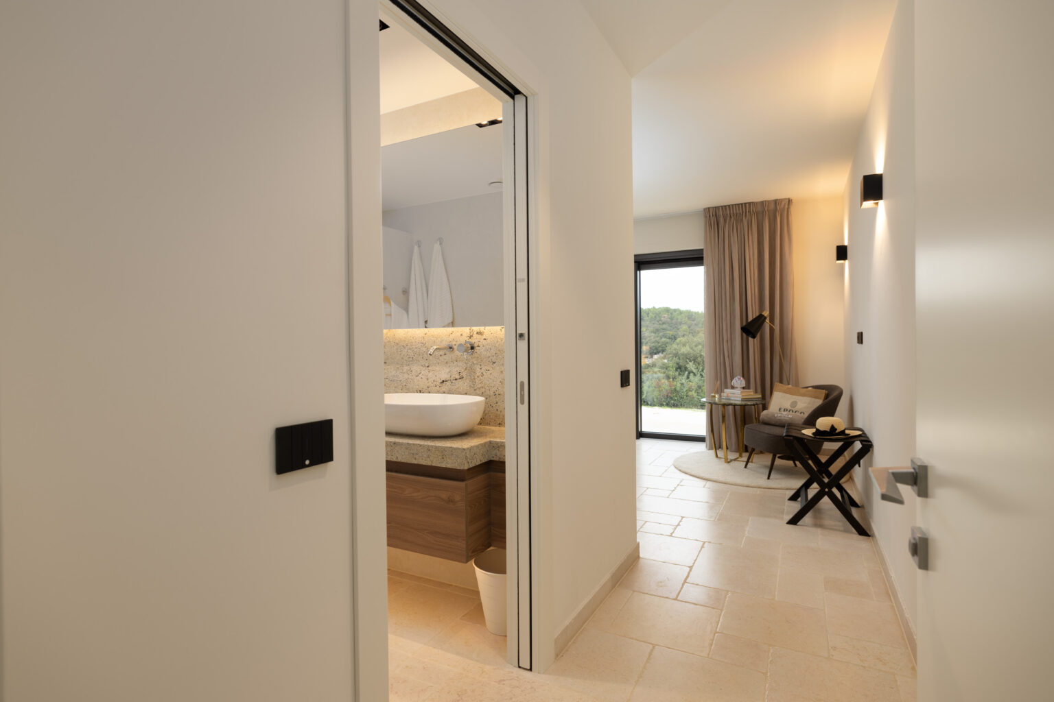 Modern hallway leading to a stylish bathroom and a cozy seating area with large windows, showcasing natural light and contemporary decor. | Only on SopranoVillas