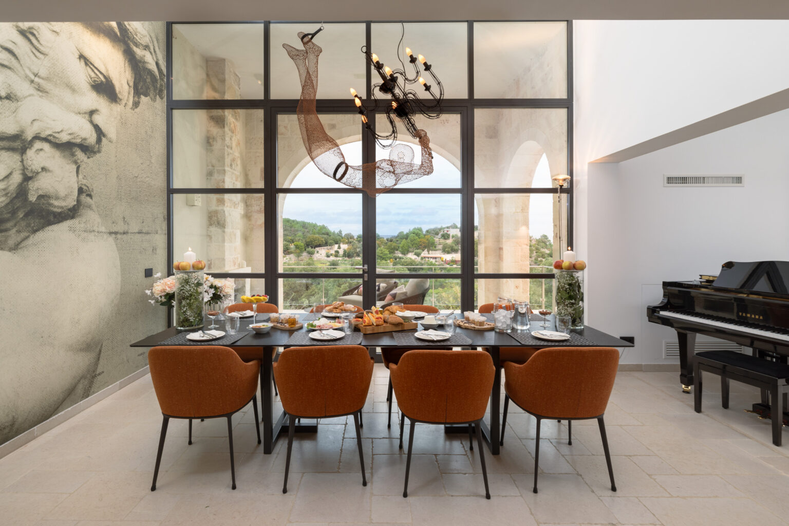 Modern dining room featuring a large table set for a meal, surrounded by stylish orange chairs. The space is illuminated by a contemporary chandelier and boasts large glass windows offering scenic outdoor views. A grand piano is positioned in the corner, enhancing the elegant ambiance. | Only on SopranoVillas