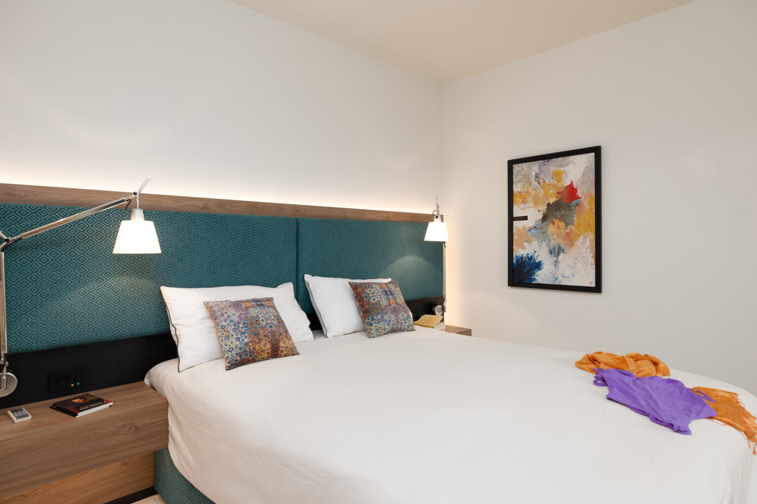 Modern hotel room featuring a comfortable double bed with decorative pillows, stylish bedside lamps, and an abstract wall art piece. The room has a clean, minimalist design with a wooden nightstand and vibrant throws. | Only on SopranoVillas