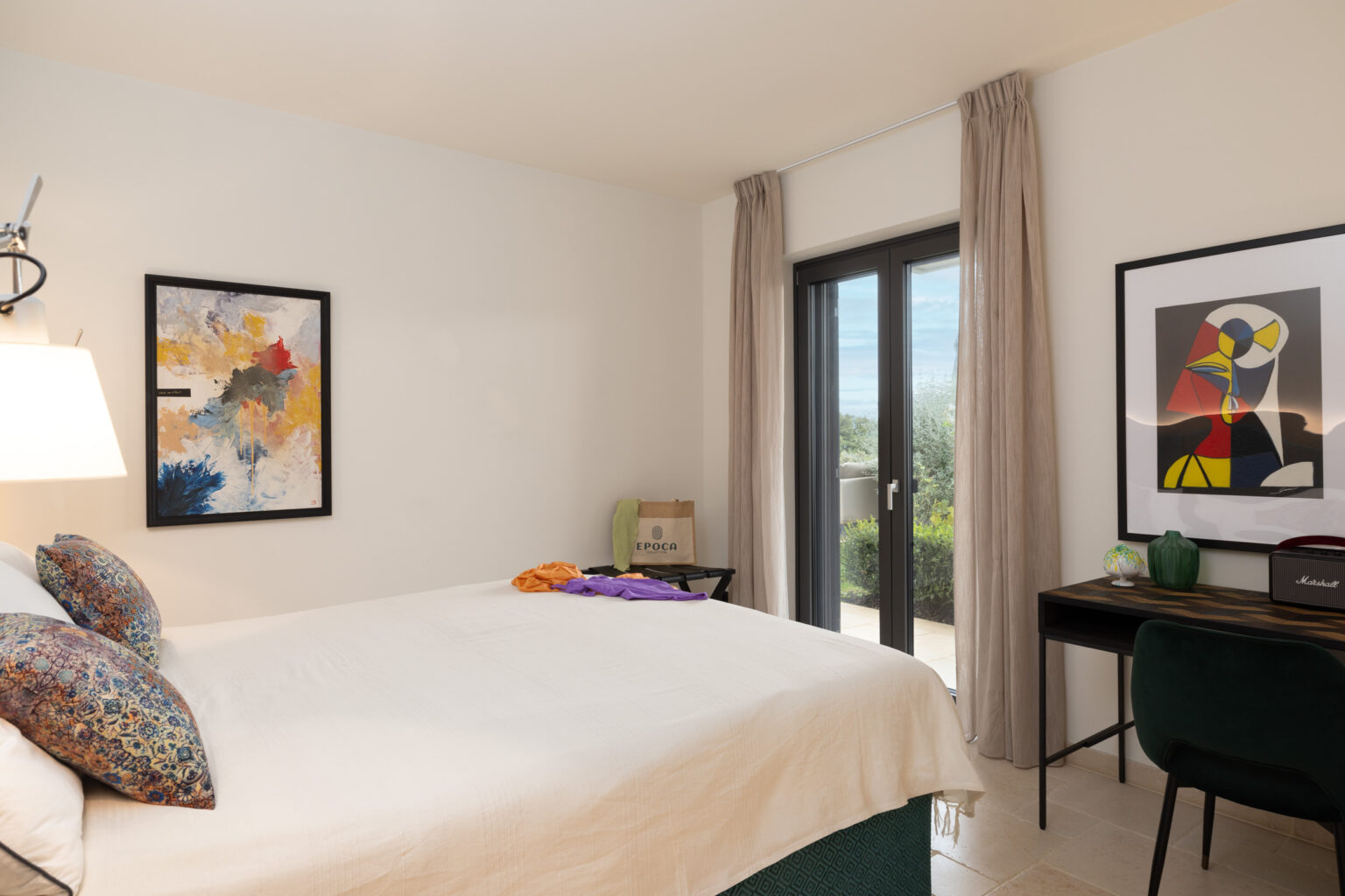 Cozy modern bedroom featuring a queen-sized bed with decorative pillows, two framed artworks on the walls, a stylish desk with a chair, and large glass doors leading to an outdoor space. | Only on SopranoVillas