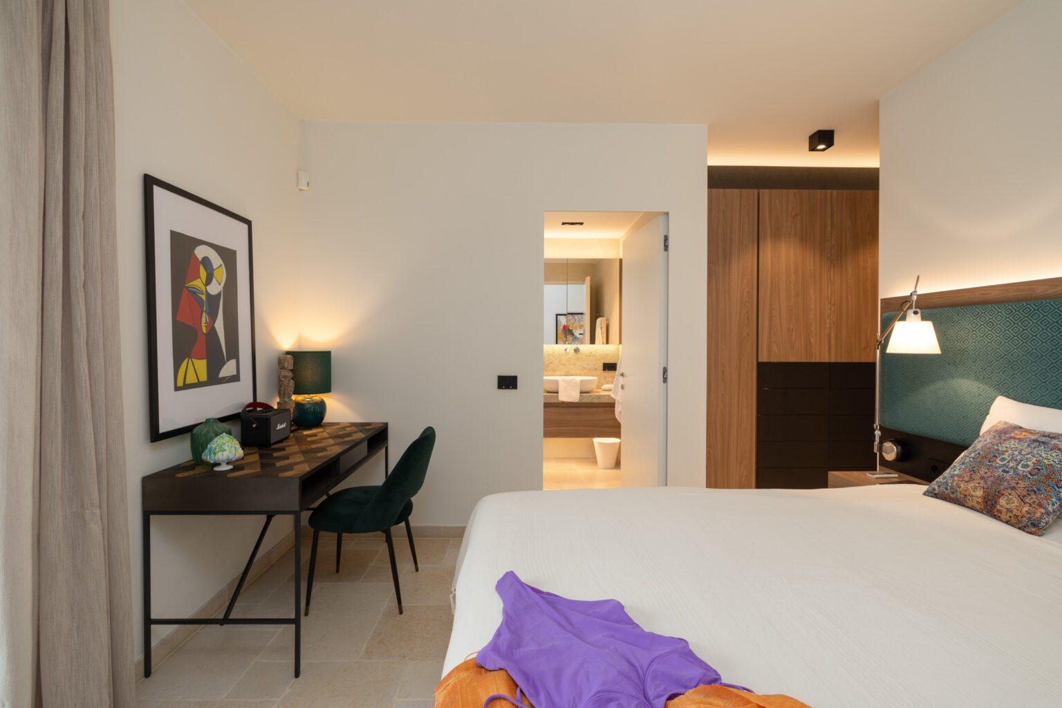 Modern hotel room featuring a comfortable bed, stylish desk with green chairs, and an en-suite bathroom. The decor includes contemporary artwork and warm lighting, creating a cozy atmosphere. | Only on SopranoVillas