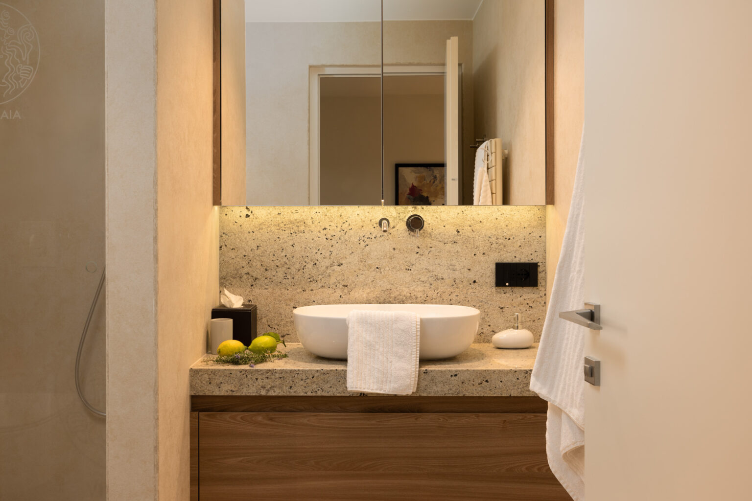 Modern bathroom design featuring a stylish bowl sink on a granite countertop, complemented by wooden cabinetry and soft lighting, creating a serene atmosphere. Fresh towels and decorative elements like limes enhance the aesthetic appeal. | Only on SopranoVillas