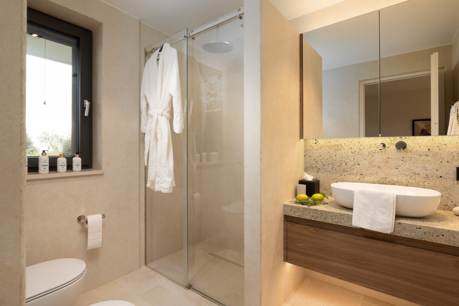 Modern bathroom featuring a glass shower, a stylish freestanding sink, and a wooden vanity. The space is adorned with a plush white bathrobe, luxury toiletries, and natural decor elements like limes, creating a serene and inviting atmosphere. | Only on SopranoVillas