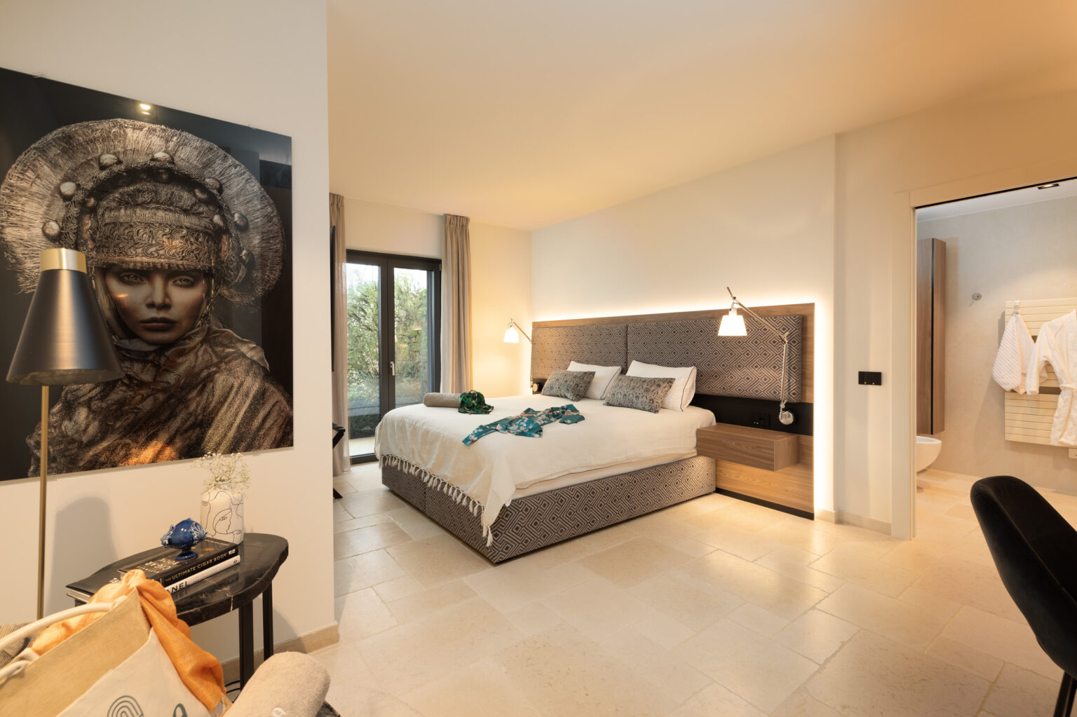 Modern hotel room featuring a king-sized bed, stylish decor, and an artistic wall portrait. The room includes natural light from large windows, a bedside table, and a bathroom entrance, creating a cozy and inviting atmosphere. | Only on SopranoVillas