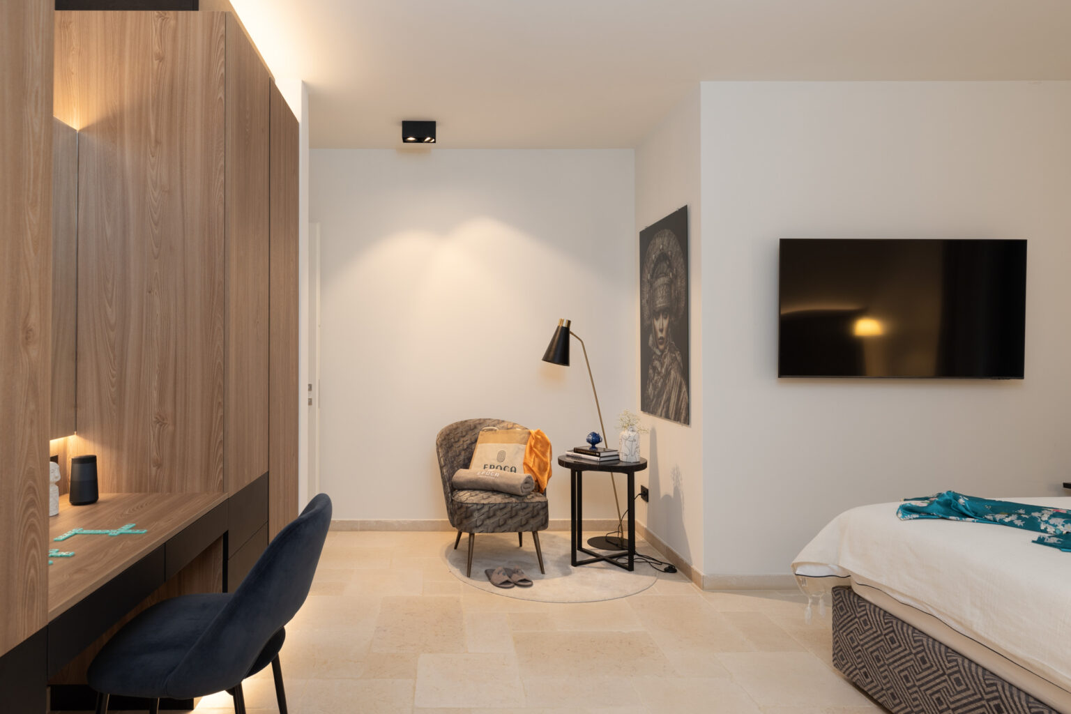 Modern hotel room featuring a cozy seating area with a patterned chair, decorative throw, and a side table. A sleek desk with a blue chair is positioned against a wooden wall, complemented by a large flat-screen TV and stylish wall art. The space is well-lit with warm lighting and showcases elegant tile flooring, creating a comfortable and inviting atmosphere. | Only on SopranoVillas