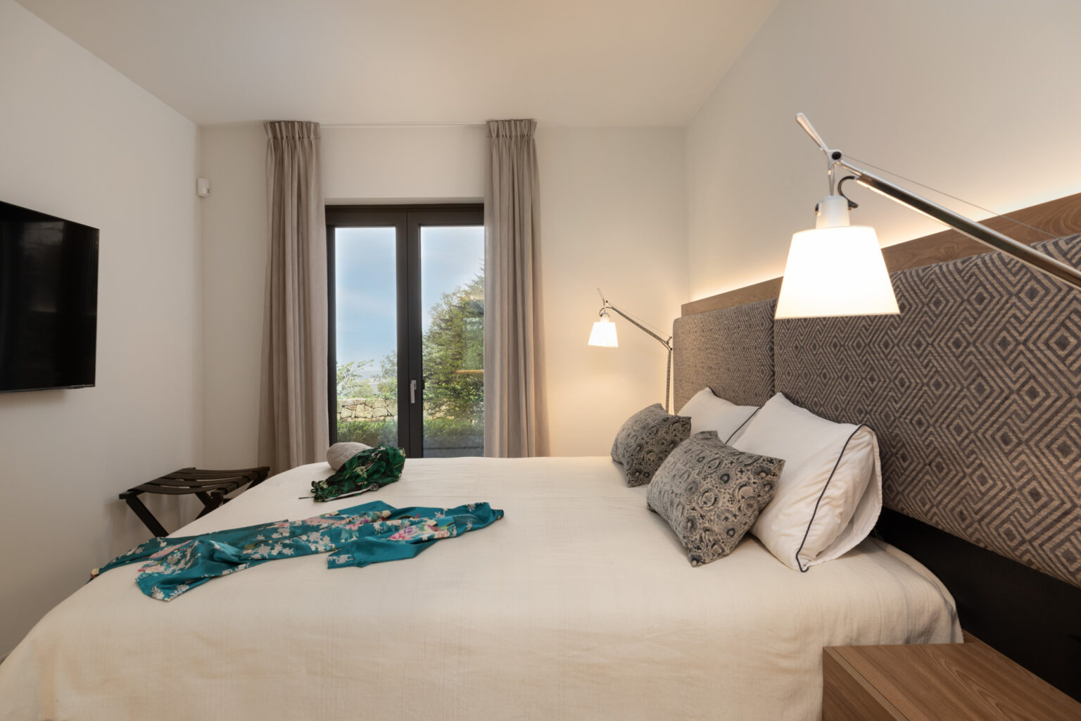 Modern bedroom with a large bed adorned with decorative pillows, a stylish bedside lamp, and a flat-screen TV. A vibrant teal garment is casually placed on the bed, alongside a hat, while large windows offer a view of greenery outside, creating a serene atmosphere. | Only on SopranoVillas