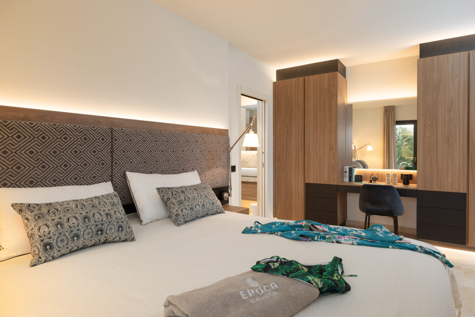 Modern hotel room featuring a comfortable bed with patterned pillows, a stylish wooden wardrobe, and a well-lit desk area. The decor includes tropical-themed accessories and soft lighting, creating a cozy ambiance for guests. | Only on SopranoVillas