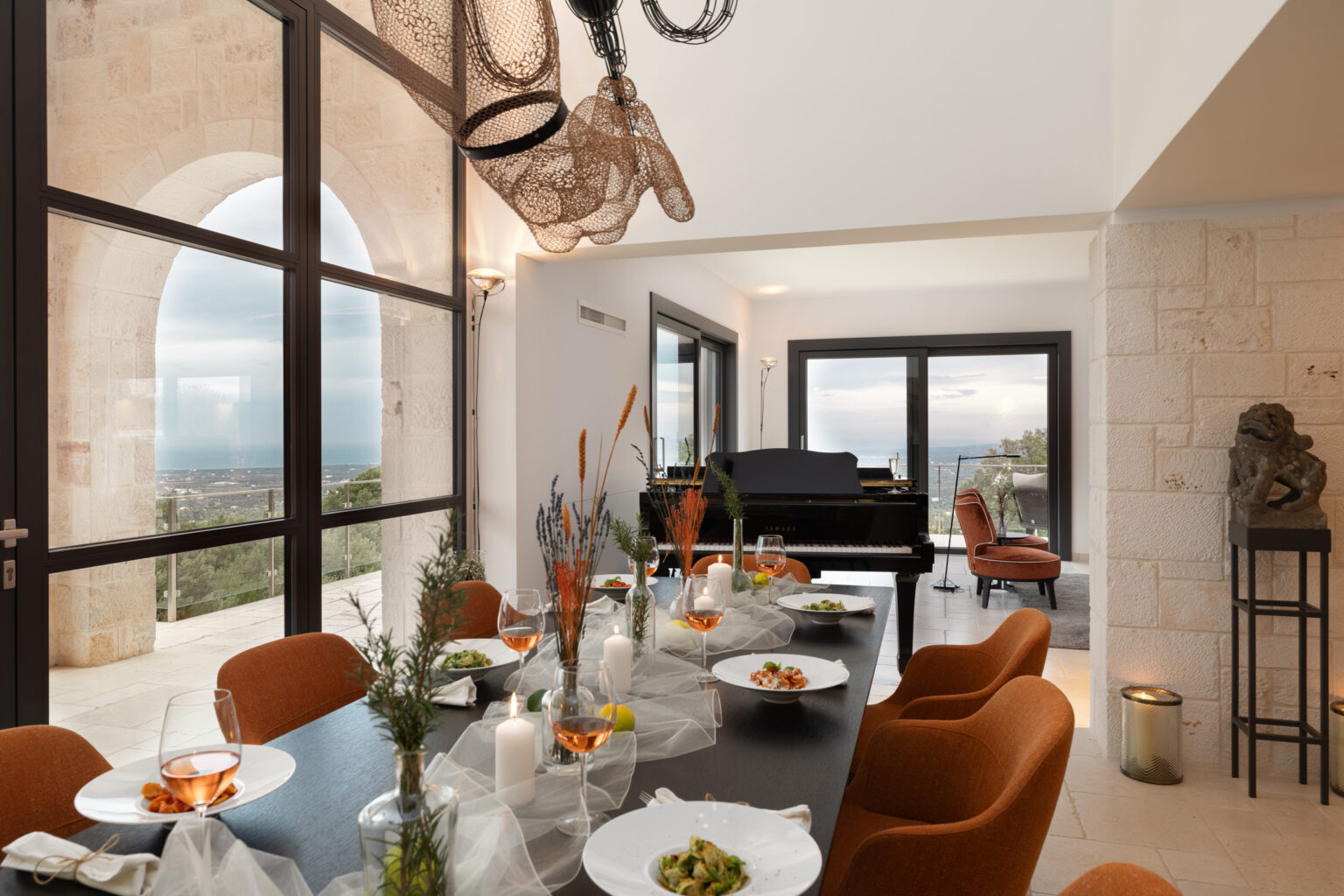 Stylish dining room featuring a long table set with gourmet dishes, elegant chairs, and a grand piano beside large windows showcasing scenic views. Modern decor elements enhance the ambiance. | Only on SopranoVillas