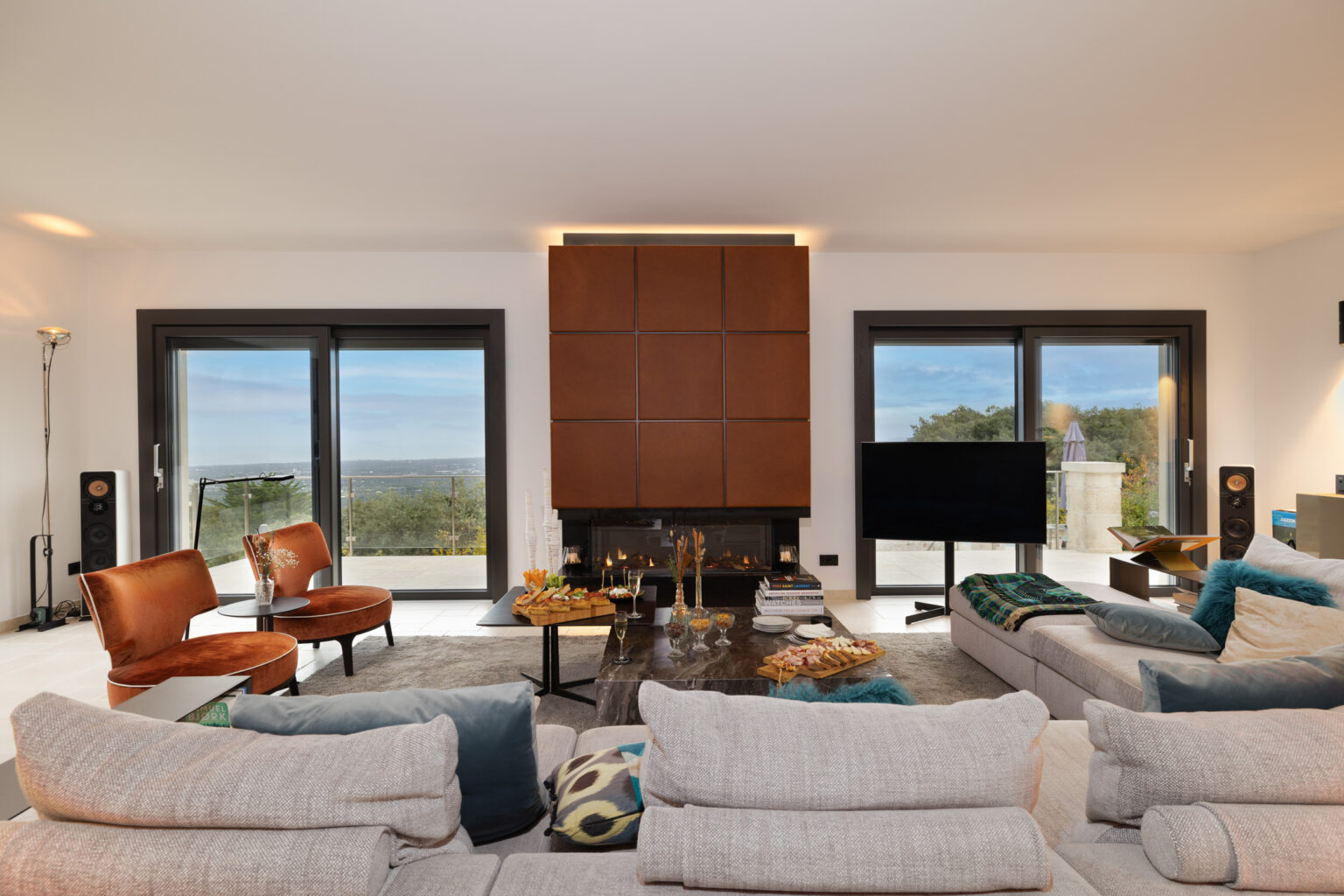 Modern living room featuring stylish gray sofas, brown accent chairs, a sleek coffee table, and large windows with scenic views. The space is elegantly decorated with contemporary art and a cozy fireplace, creating a warm and inviting atmosphere. | Only on SopranoVillas